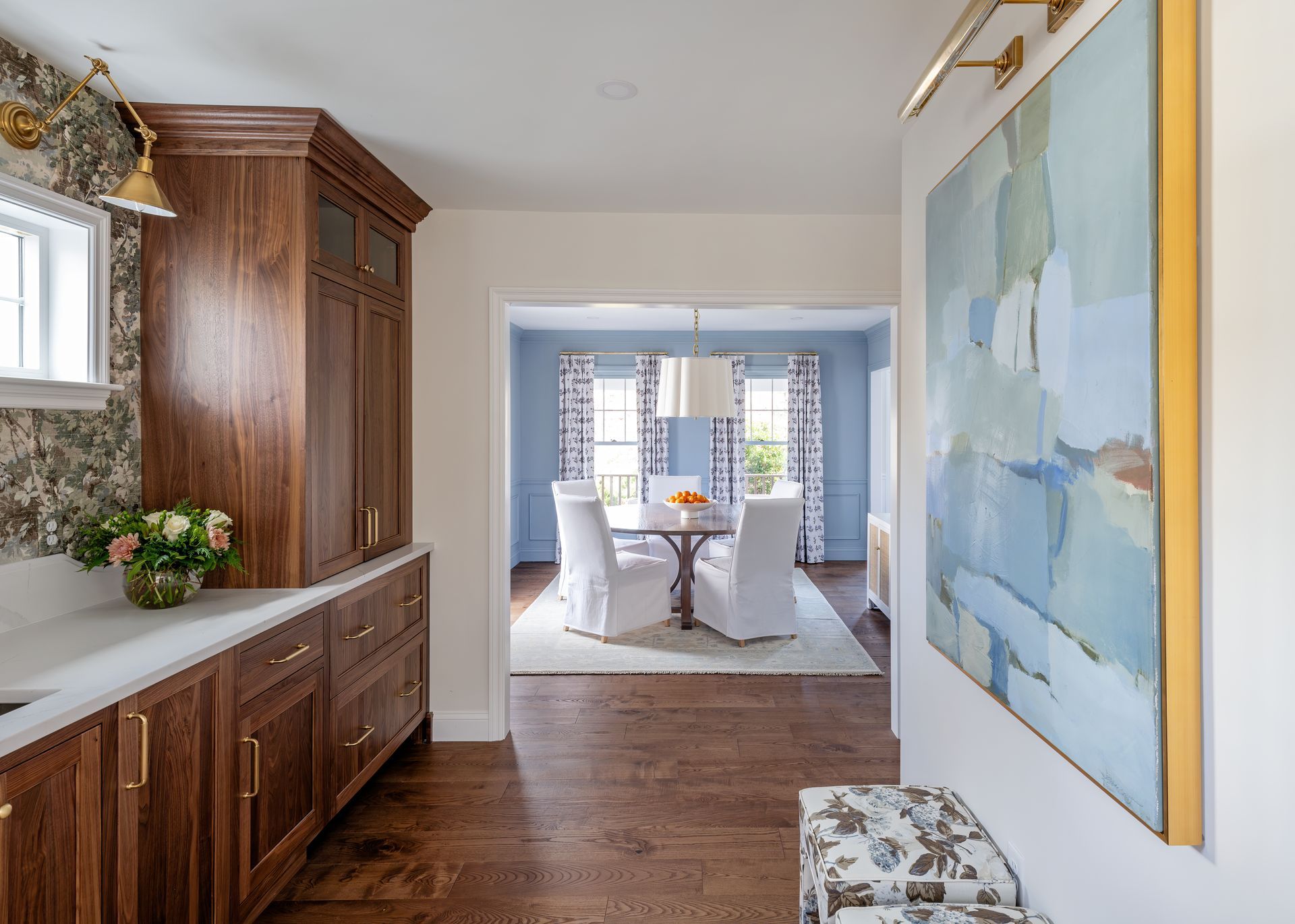 Hallway with dark wood sideboard, floral bench, and blue abstract art leading to a bright dining room
