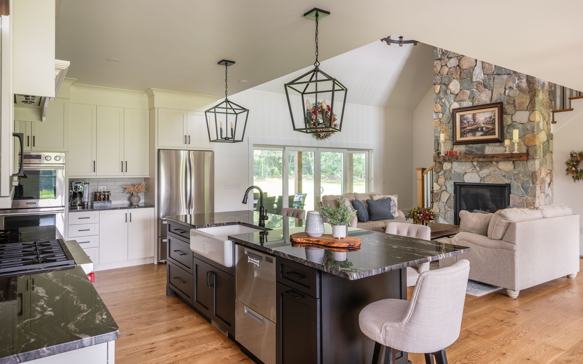 Modern kitchen with island, bar stools, and open view into a living room with stone fireplace.