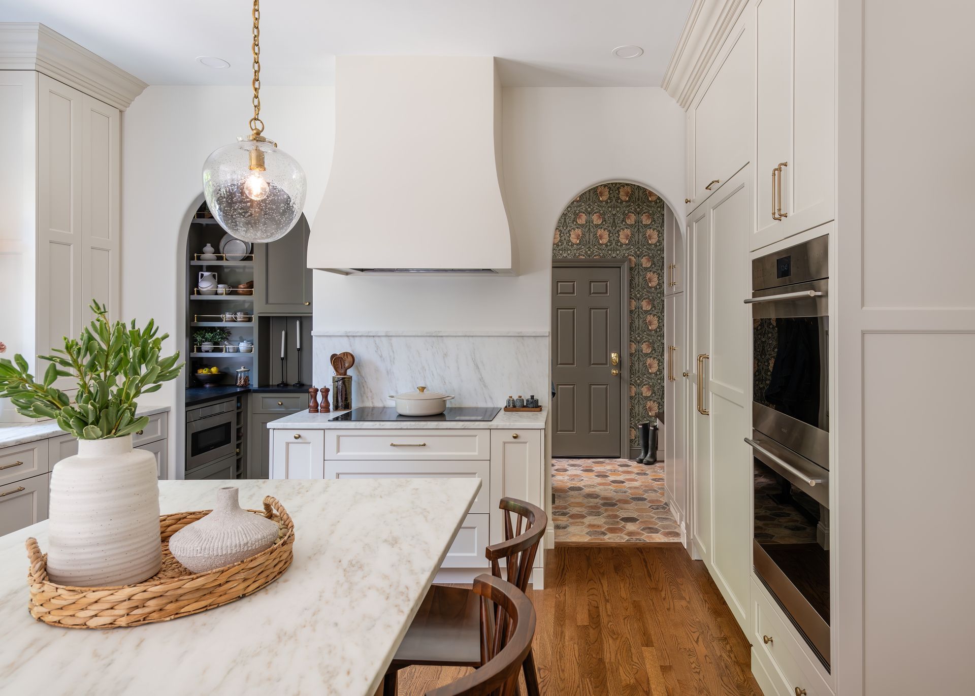 Bright white kitchen with marble island, woven baskets, wood floors, and arched doorway.