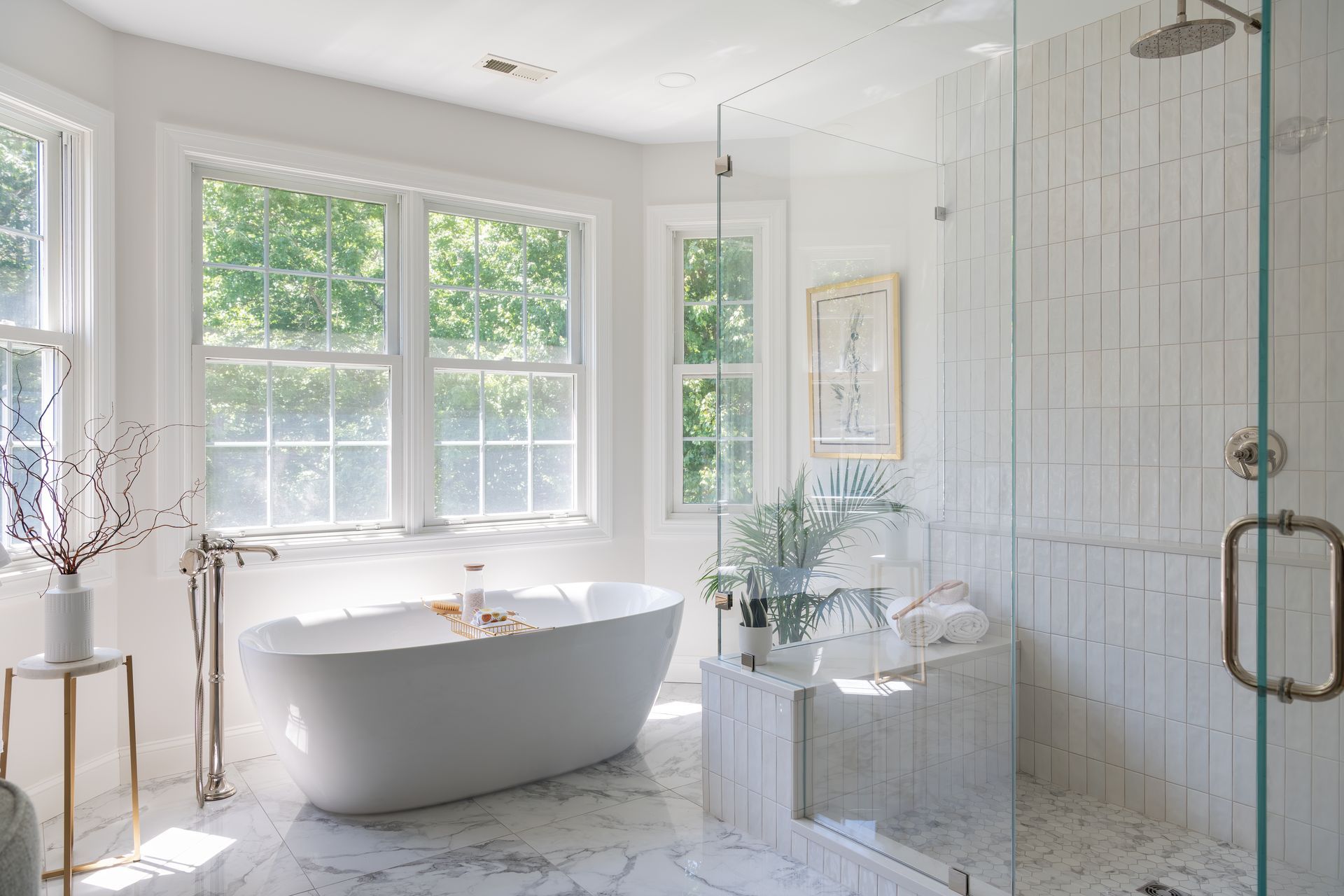 Bright white bathroom with a freestanding tub, glass shower, and large windows overlooking greenery