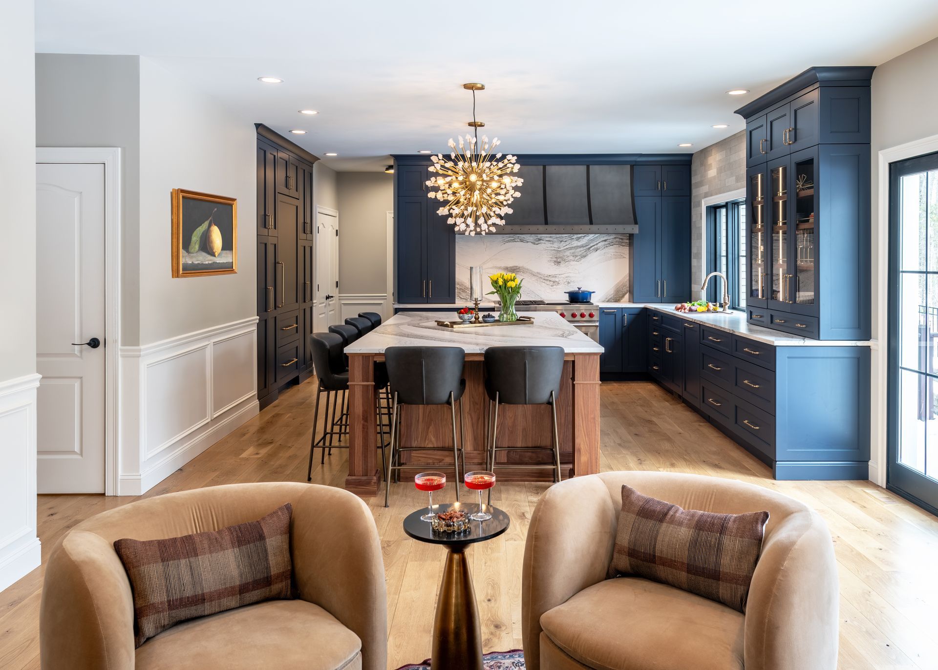 Modern kitchen with navy cabinets, marble island, brass chandelier, and two beige chairs in the foreground