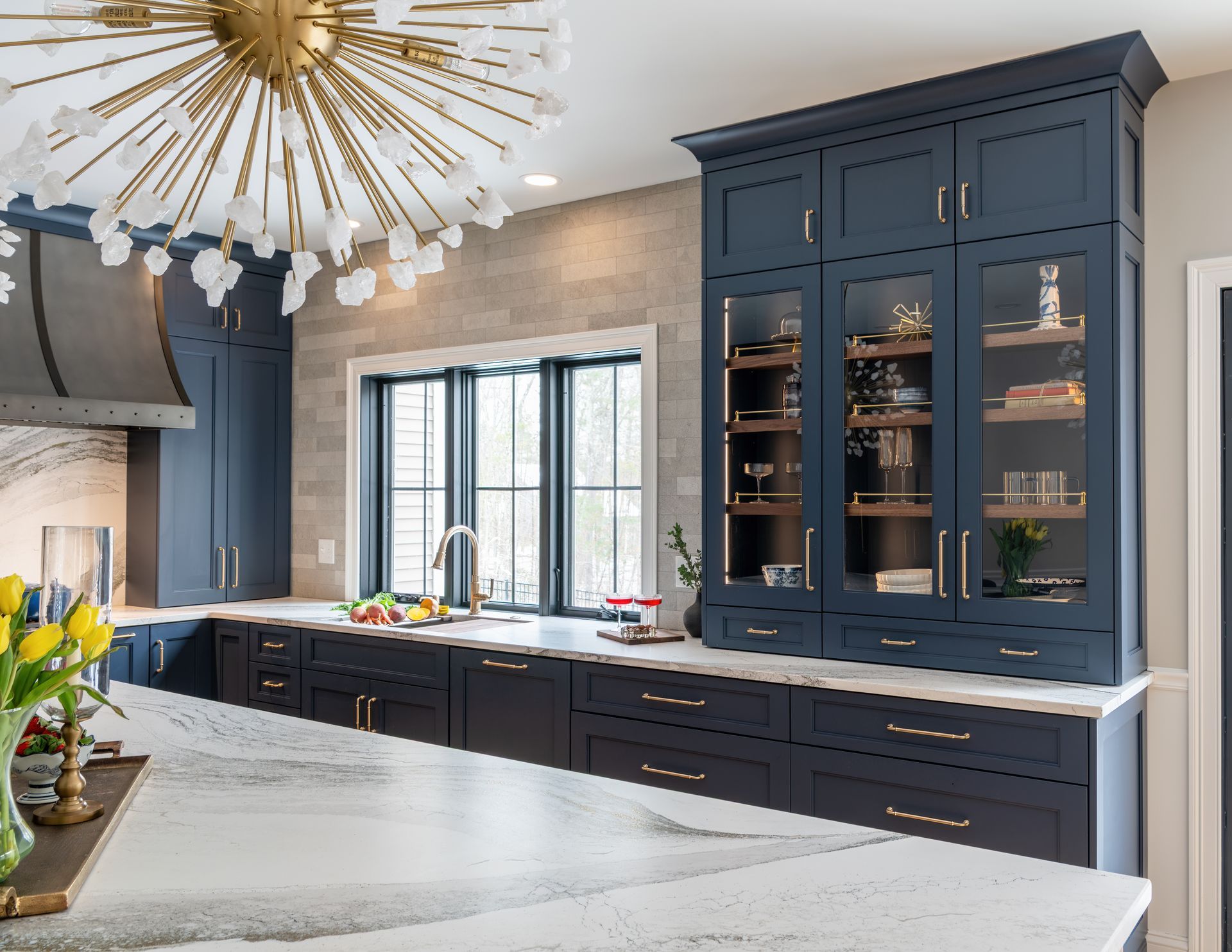 Modern kitchen with navy cabinets, marble island, brass chandelier, and large window above the sink