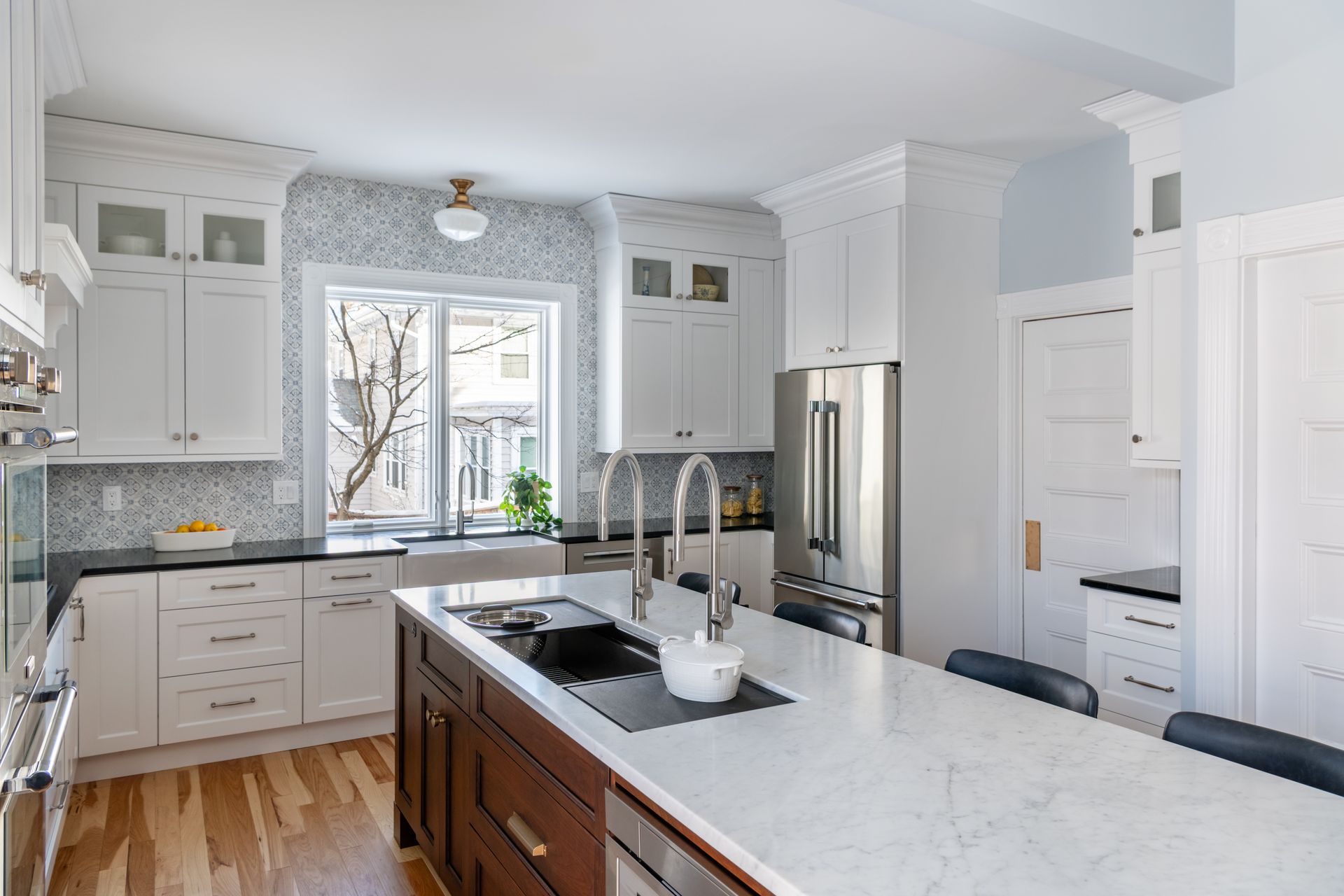 Bright modern kitchen with white cabinets, marble island, stainless fridge, and large window.