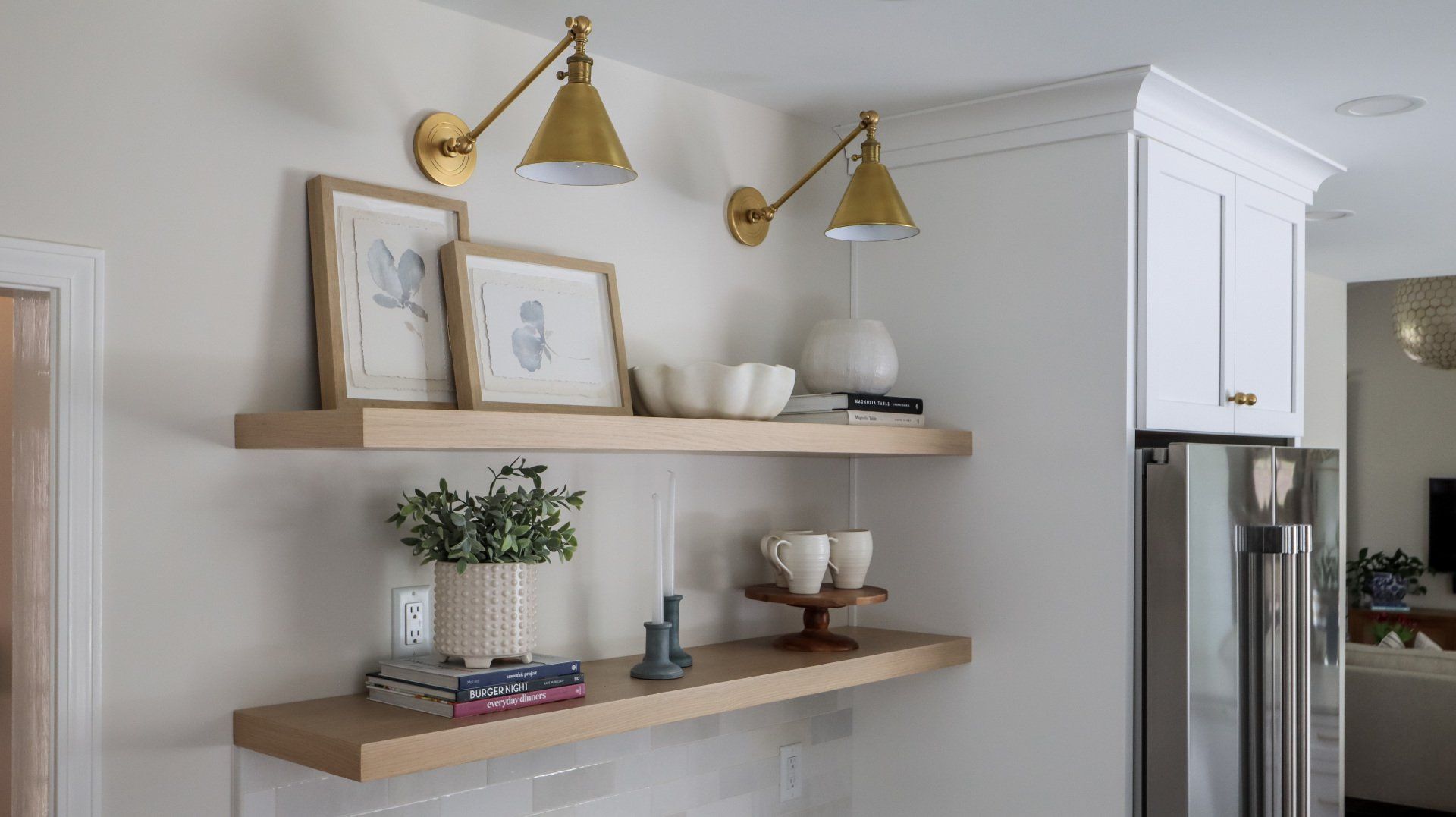 Kitchen wall with floating shelves, framed art, plants, bowls, and brass sconces next to a stainless fridge