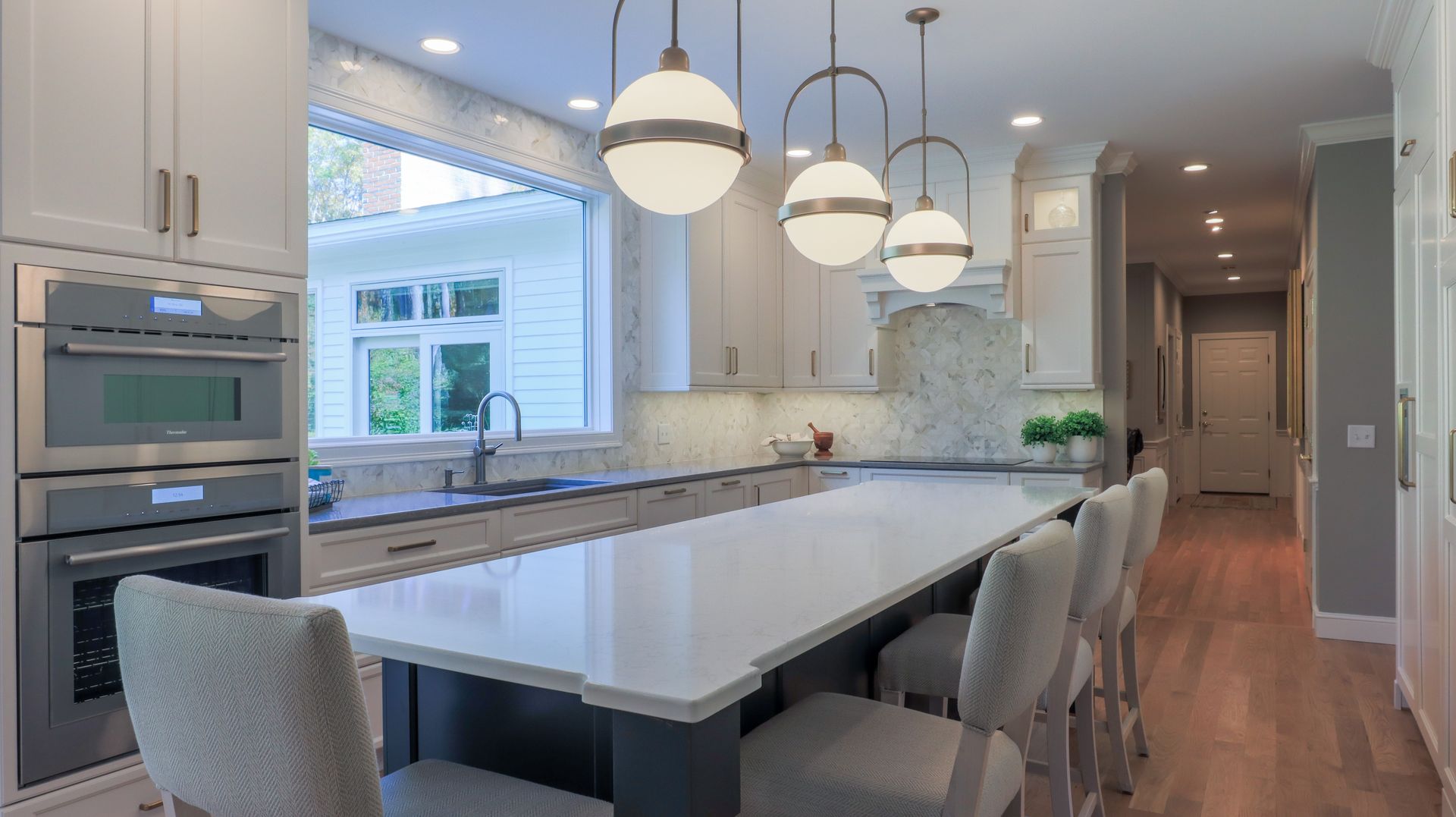 Modern white kitchen with large island, pendant lights, built-in ovens, and bright window seating area