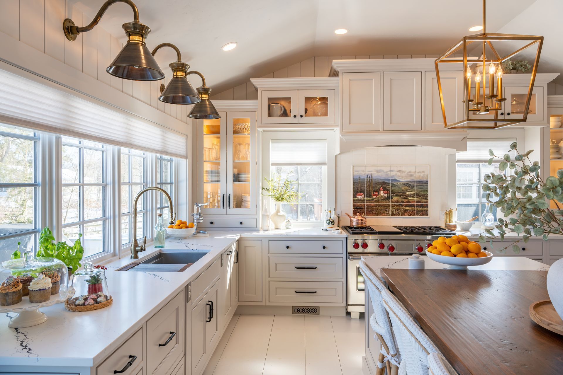 Bright, modern kitchen with white cabinets, large windows, brass lights, and a wooden island with fruit.