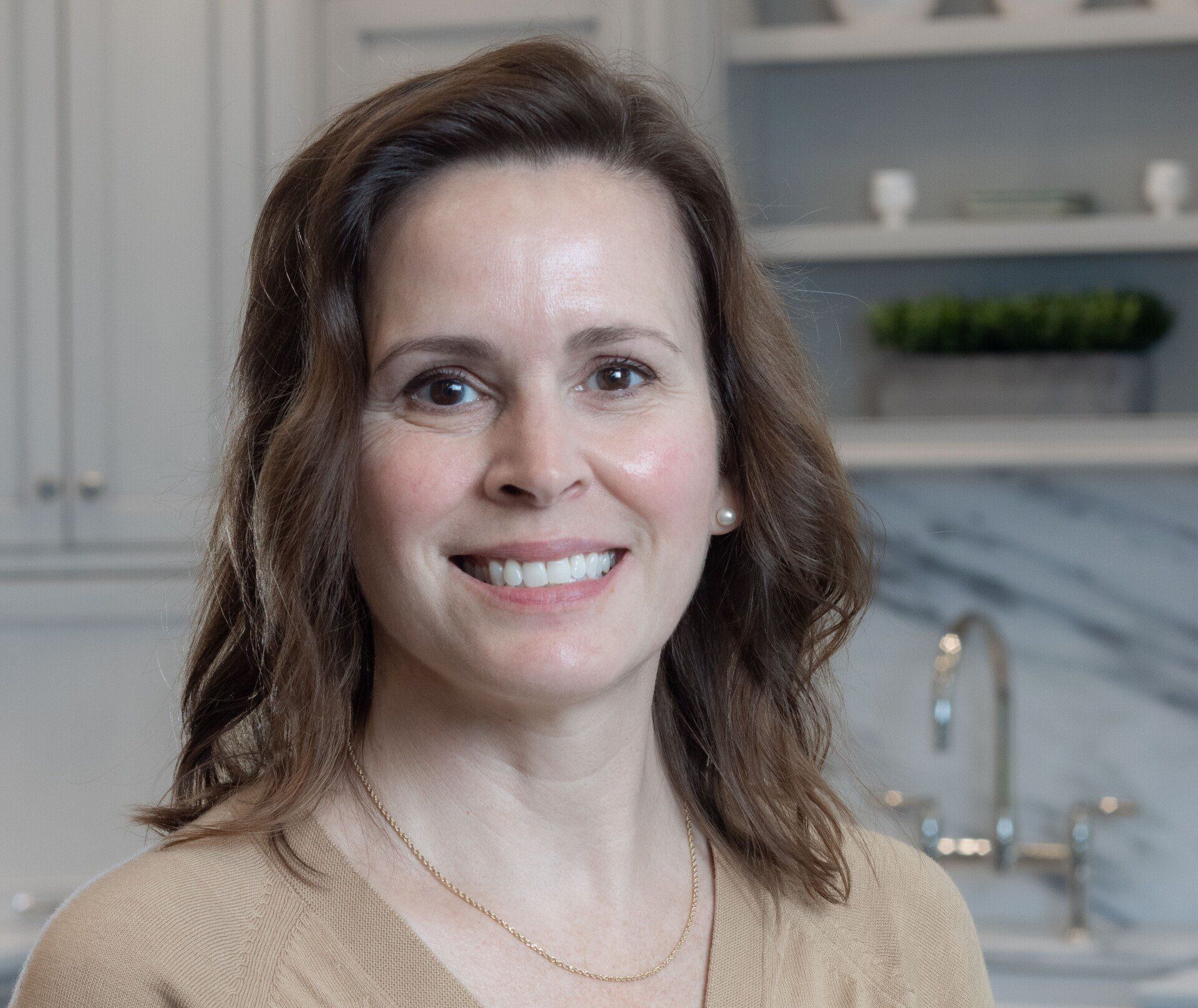 Smiling woman in a beige sweater standing in a bright kitchen with marble backsplash