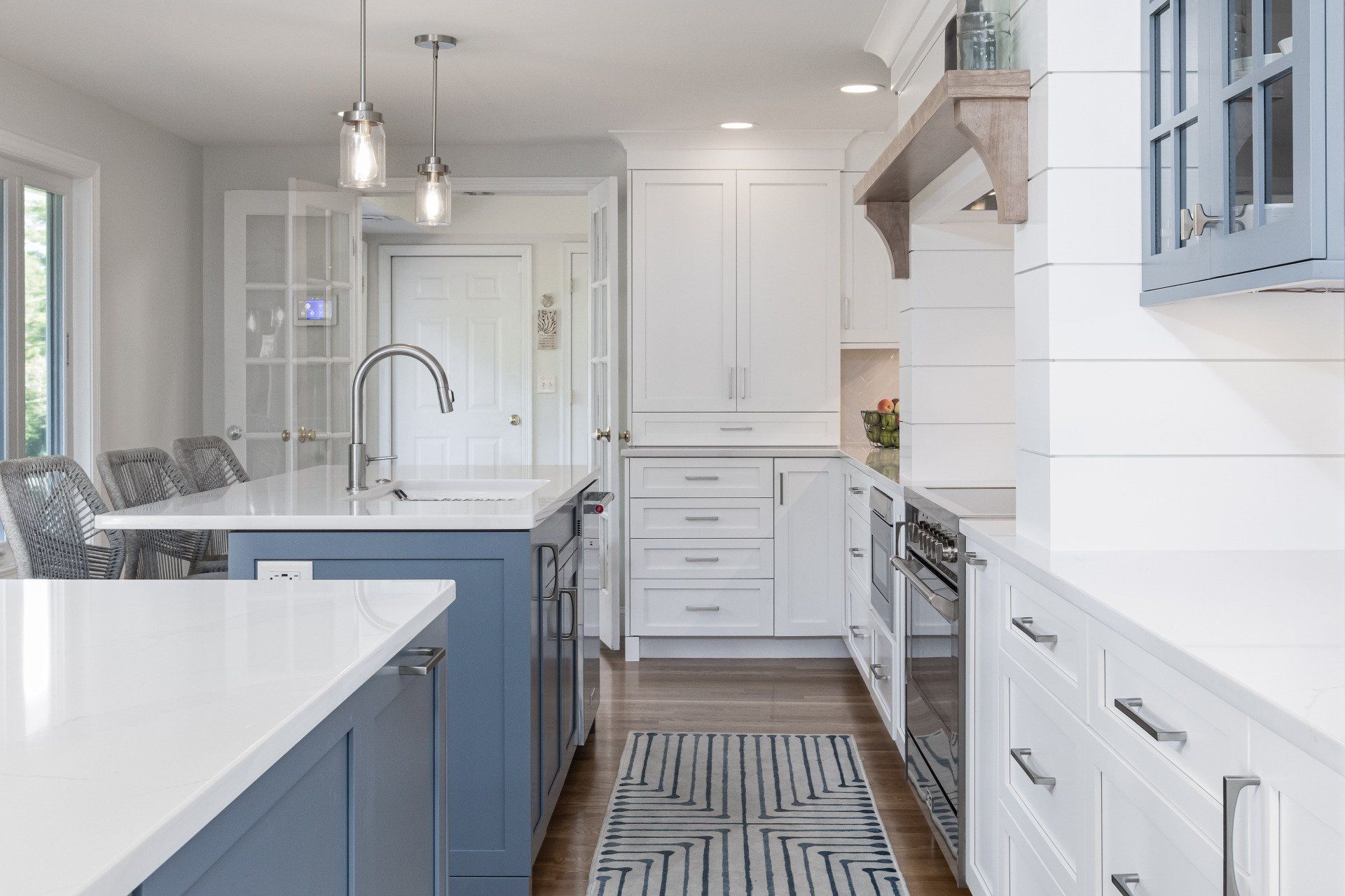 Bright white kitchen with blue island cabinets, farmhouse sink, pendant lights, and striped runner rug.