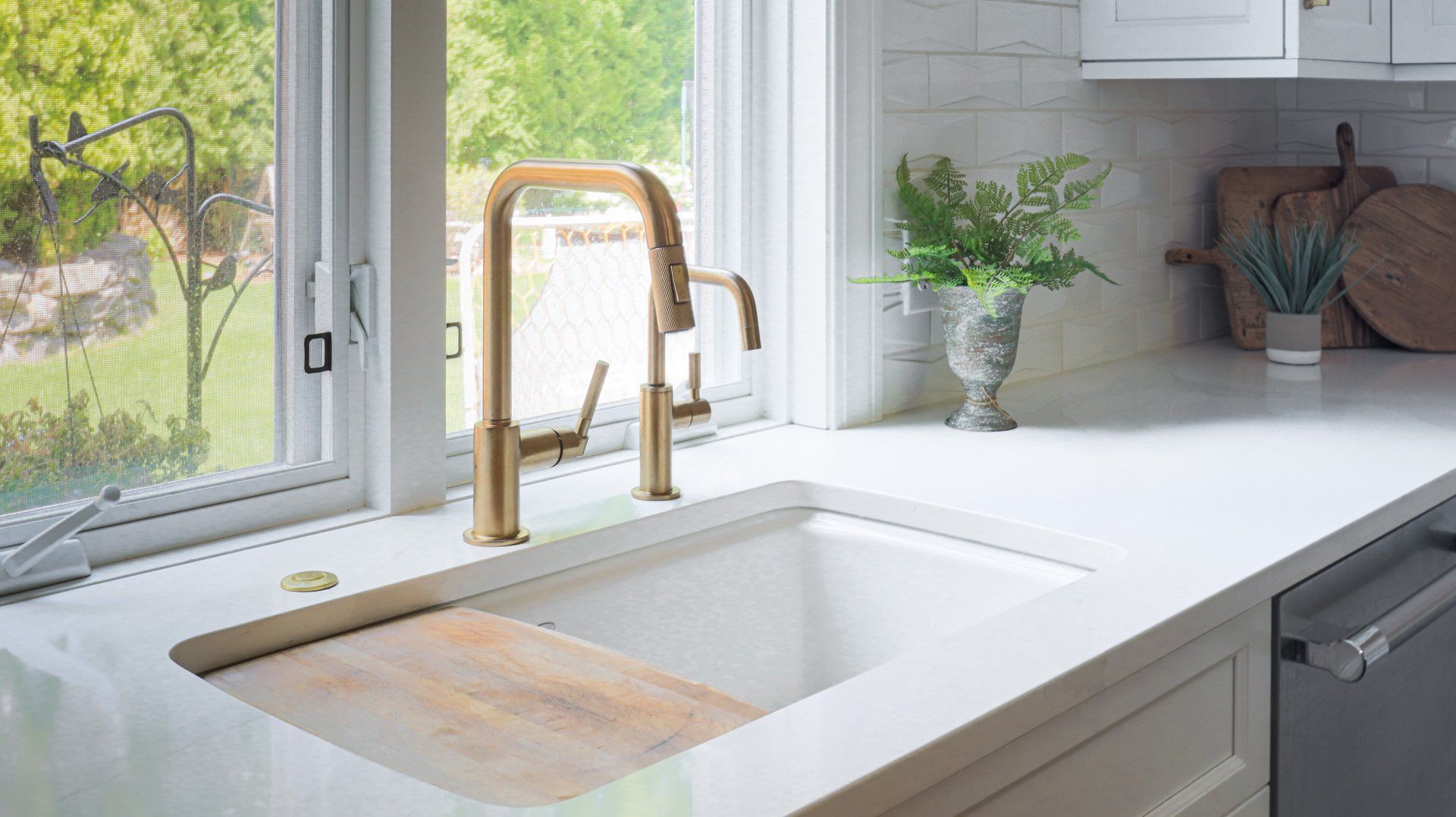 Bright kitchen sink with brass faucet, white countertop, and window view of green trees
