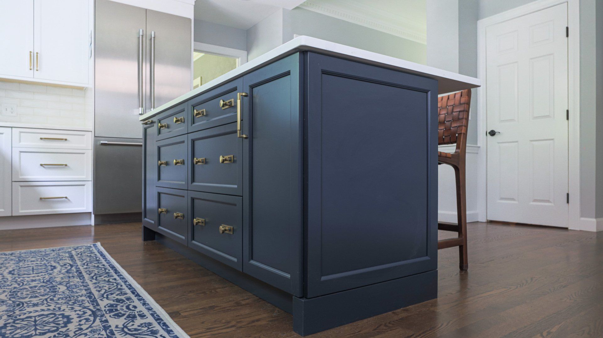 Modern kitchen with a dark blue island, stainless refrigerator, white cabinets, and hardwood floor.