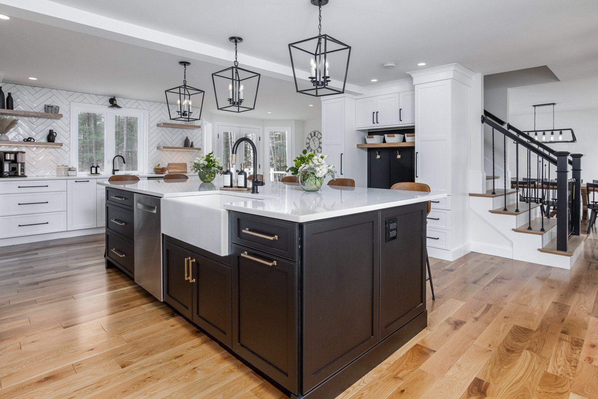 Modern white kitchen with dark island, pendant lights, hardwood floors, and staircase in the background