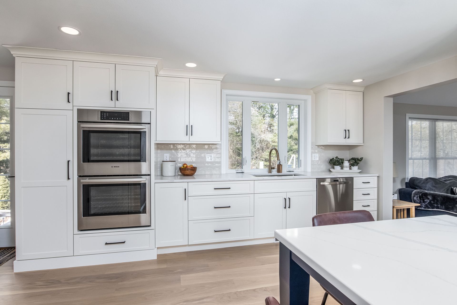 Bright modern white kitchen with built-in ovens, island, and windows overlooking a backyard