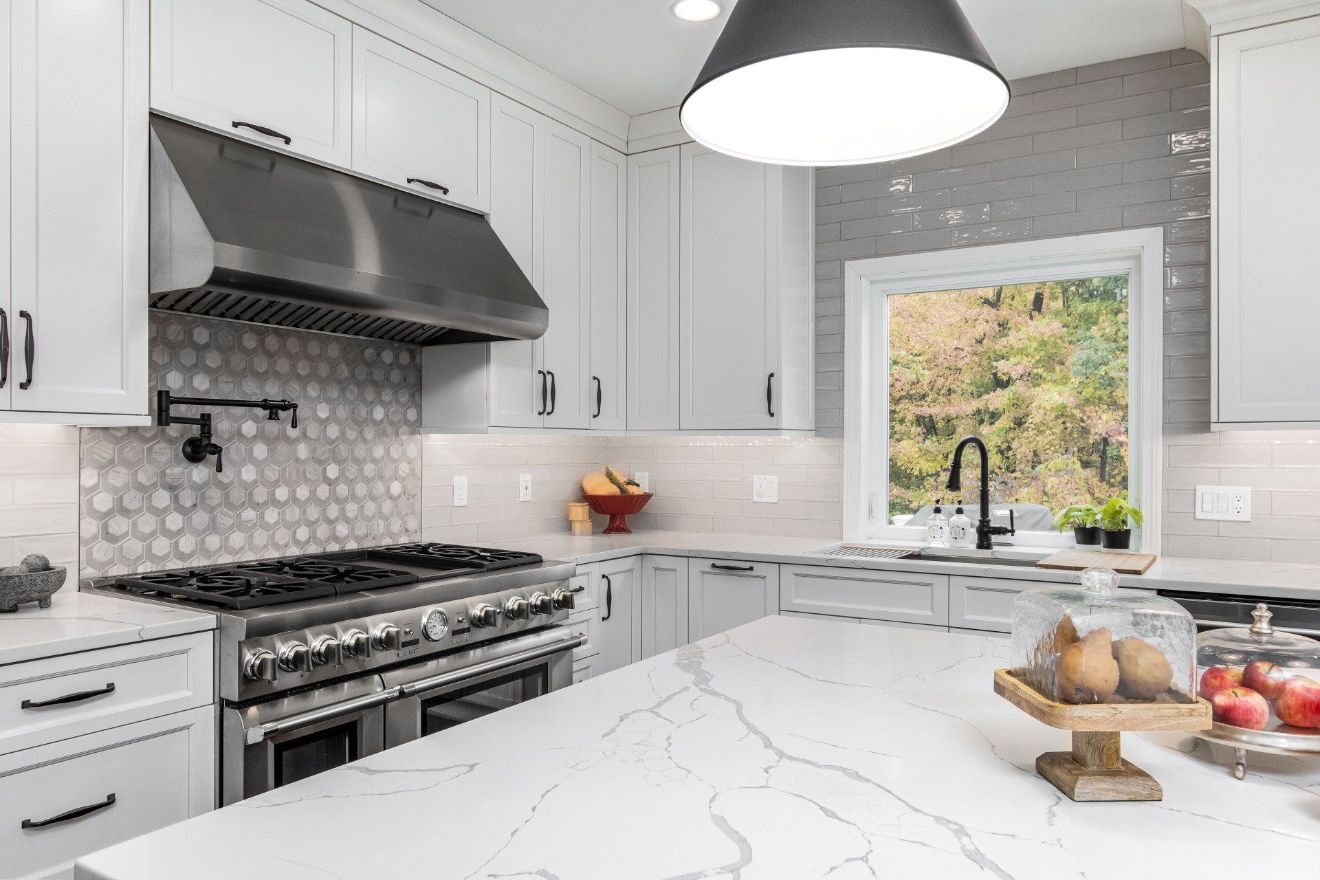 Modern white kitchen with marble island, stainless steel stove, pendant light, and window view of greenery