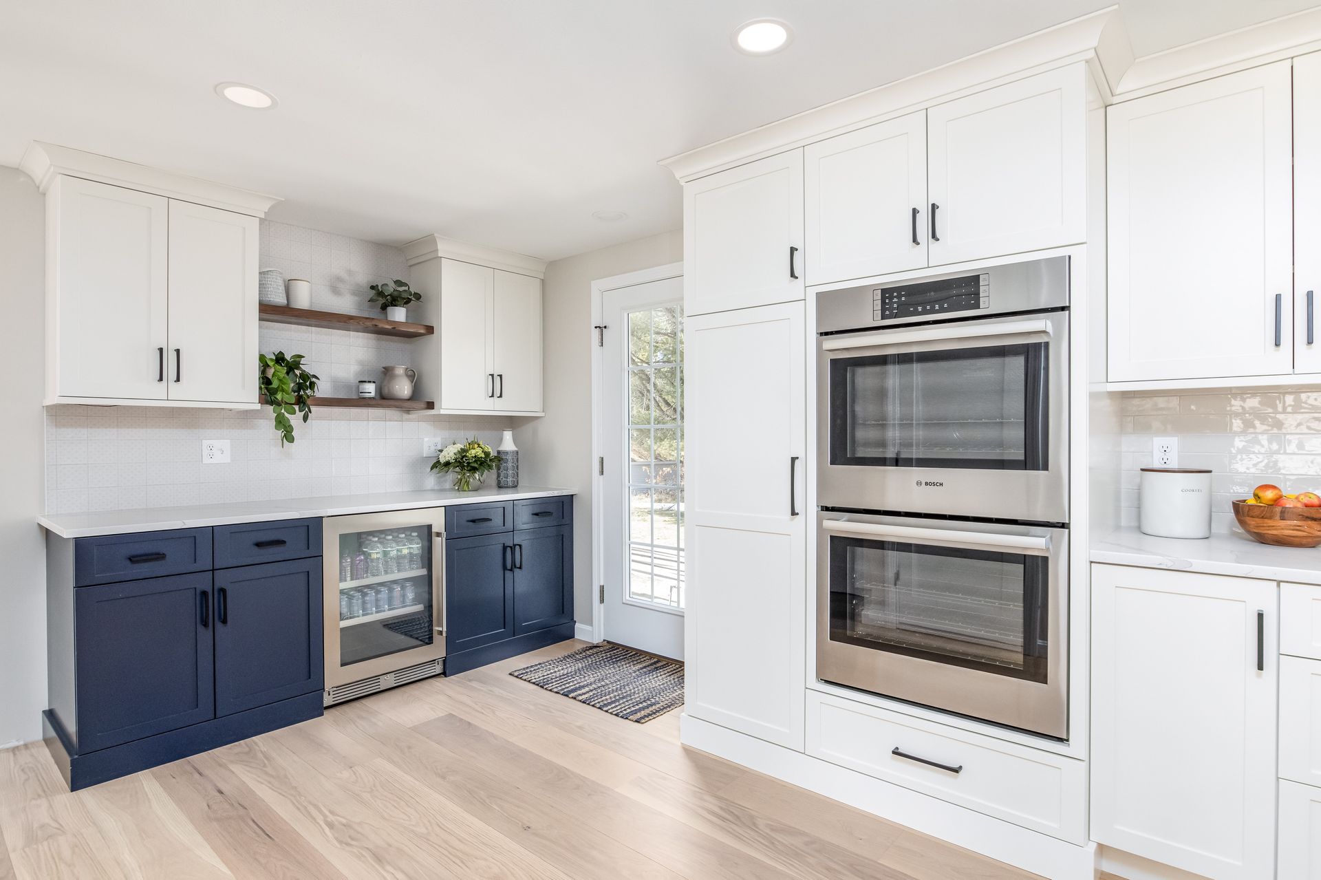 Bright white kitchen with navy lower cabinets, stainless appliances, and built-in double ovens