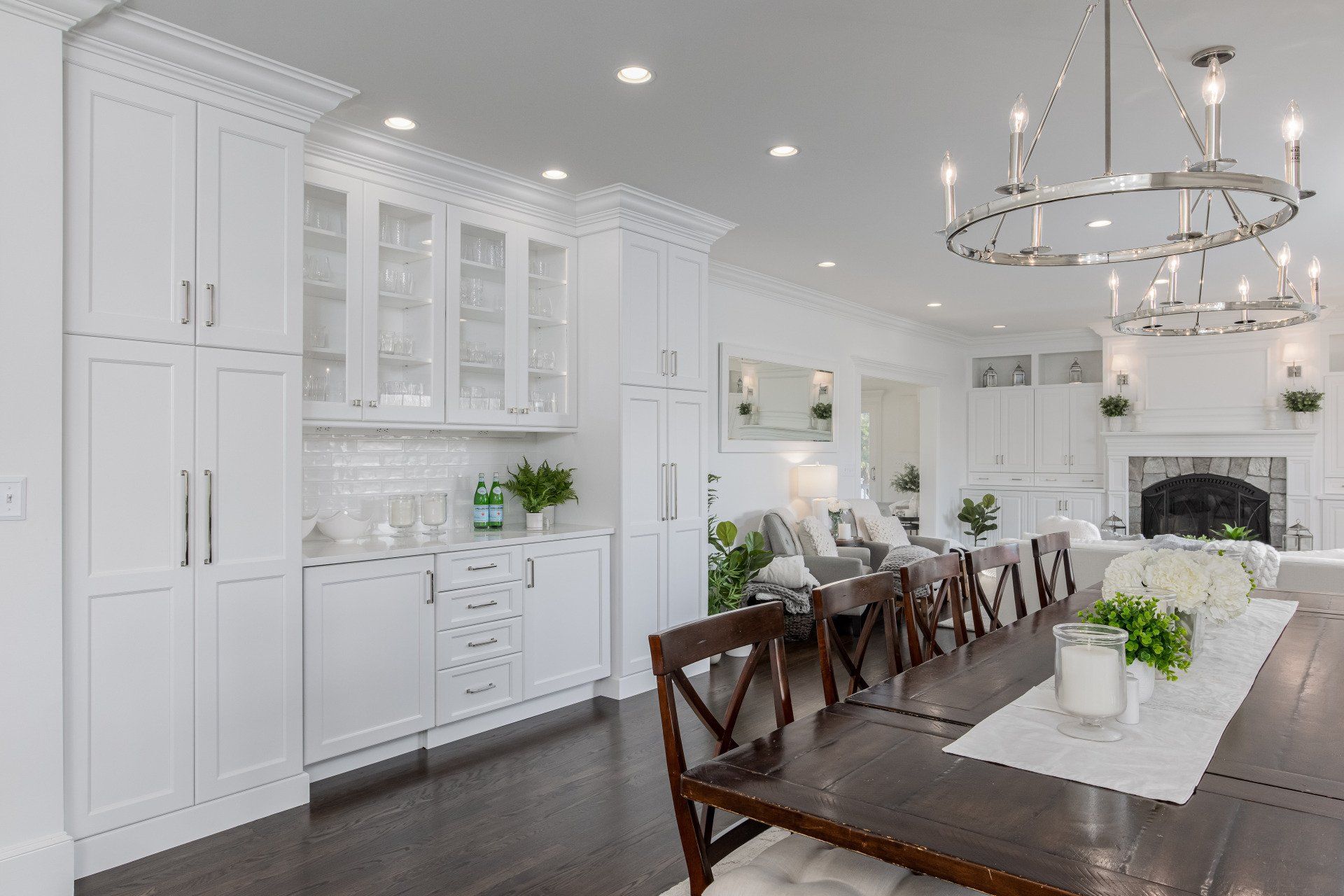 Bright white modern dining room and kitchen with a long wooden table, chandelier, and fireplace.