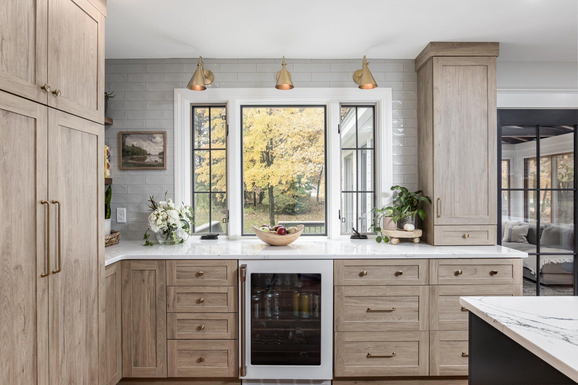 Bright kitchen with light wood cabinets, large window, white countertops, and small plants.