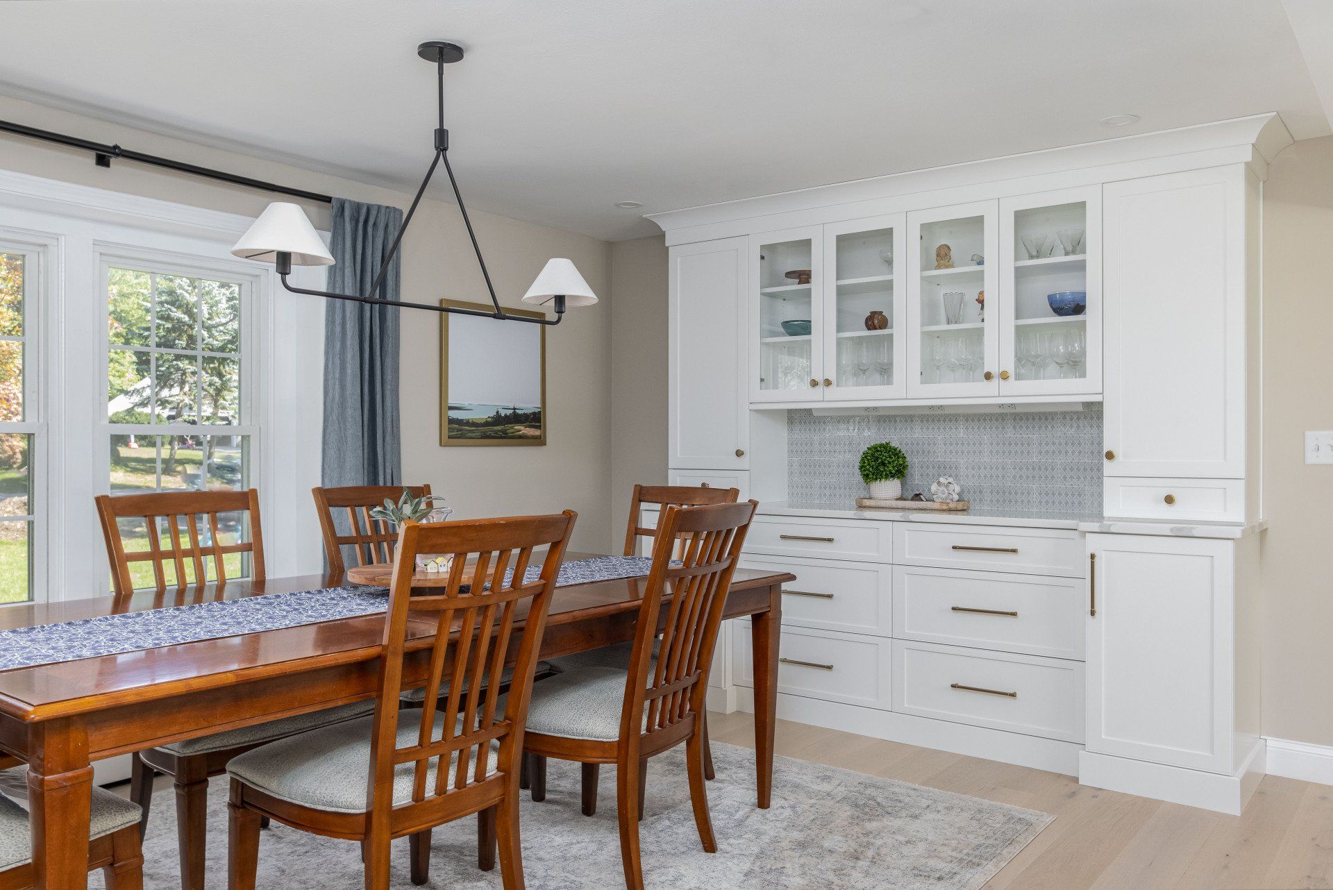 Bright dining room with wooden table, chairs, white cabinets, and large windows with blue curtains