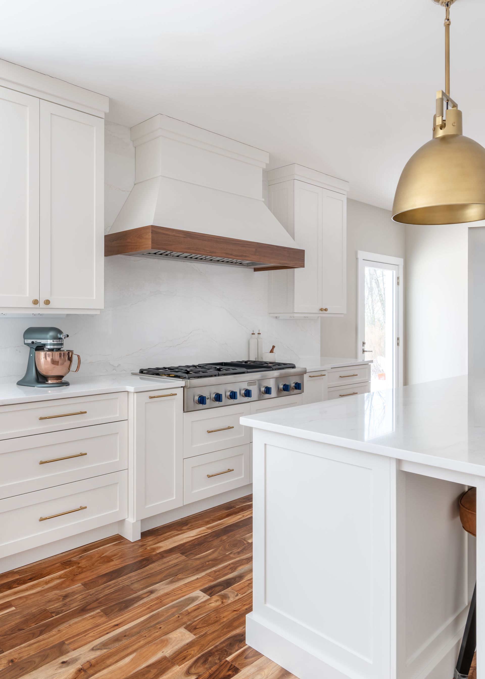 Bright white kitchen with gold pendant light, white cabinets, wood floor, and gas range with wood hood.