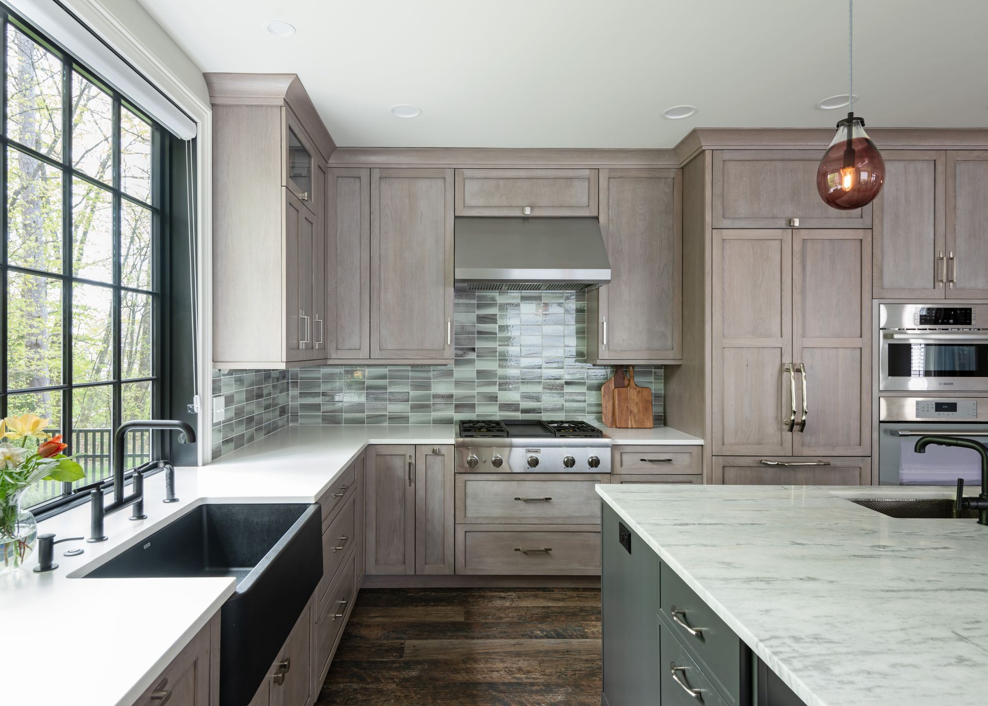 Modern kitchen with gray cabinets, marble island, farmhouse sink, and large window above black countertops