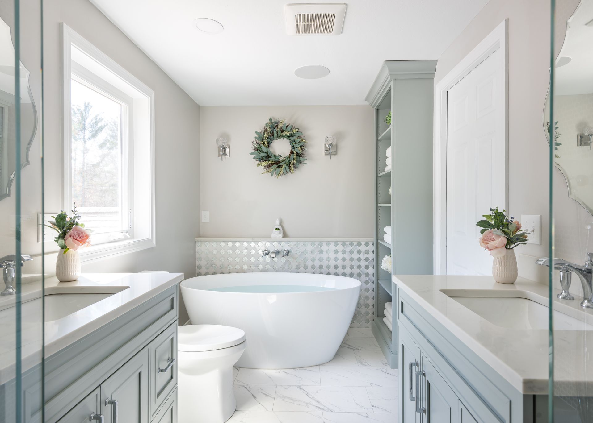 Bright white bathroom with freestanding tub, mint cabinetry, marble counters, and wreath wall decor.