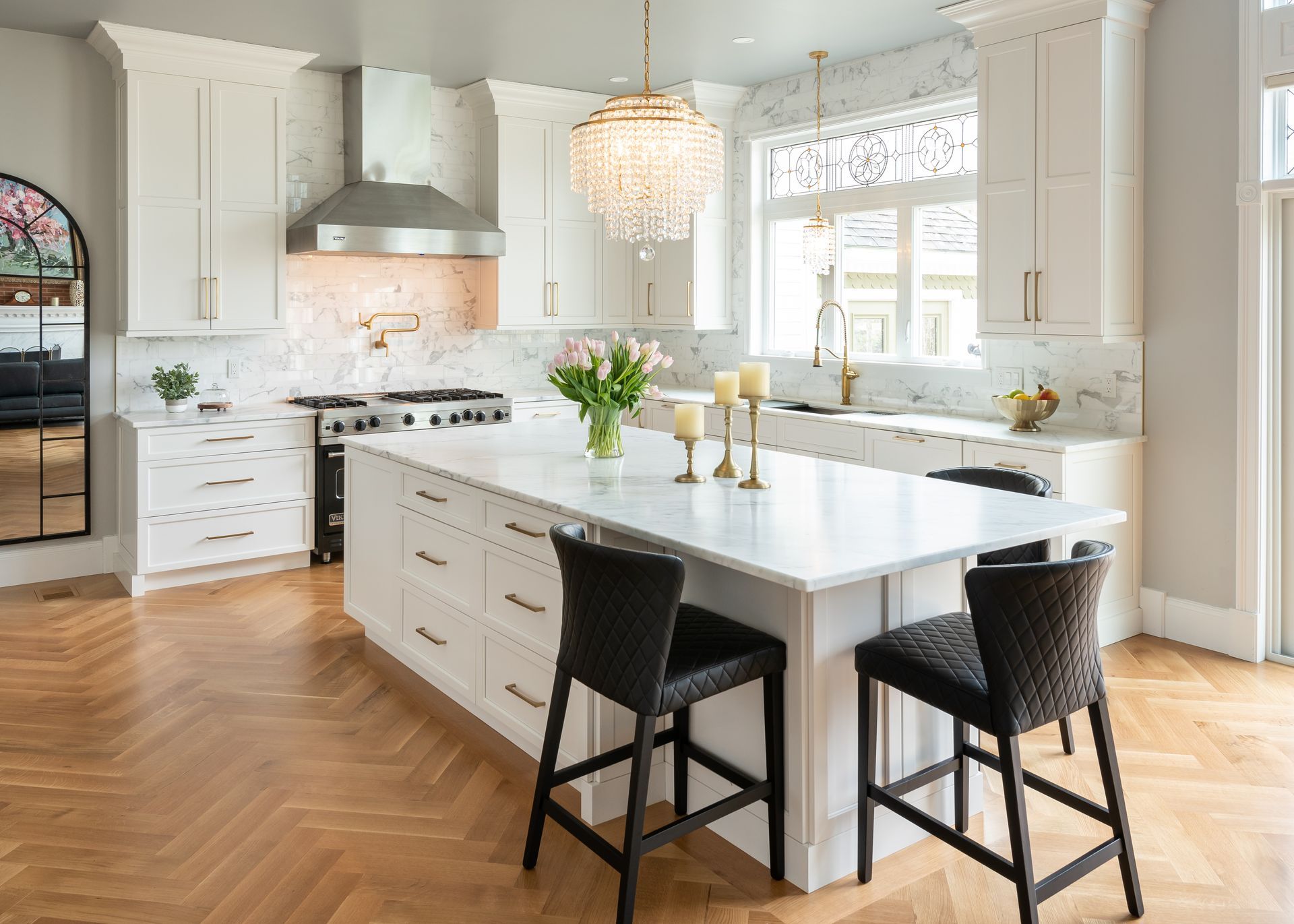 Bright white kitchen with large island, black stools, stainless range hood, and sunlit windows