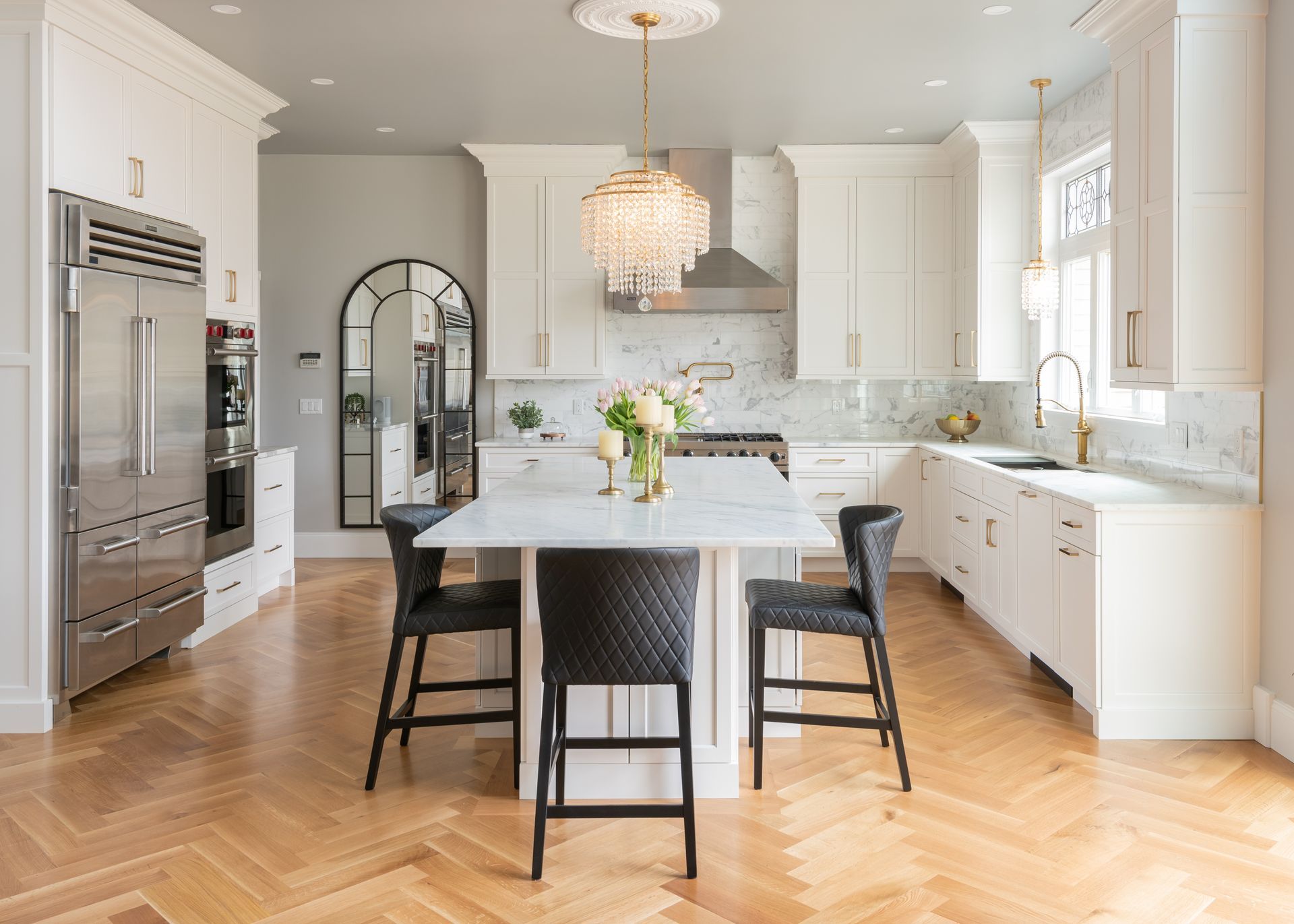 Bright white kitchen with marble island, black barstools, stainless appliances, and herringbone wood floor.
