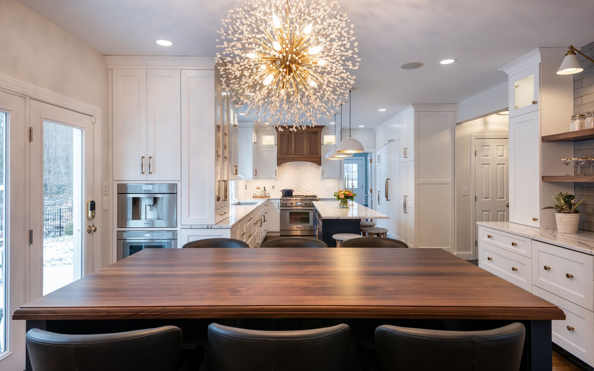 Bright white kitchen with a wooden island table and a sparkling chandelier above.