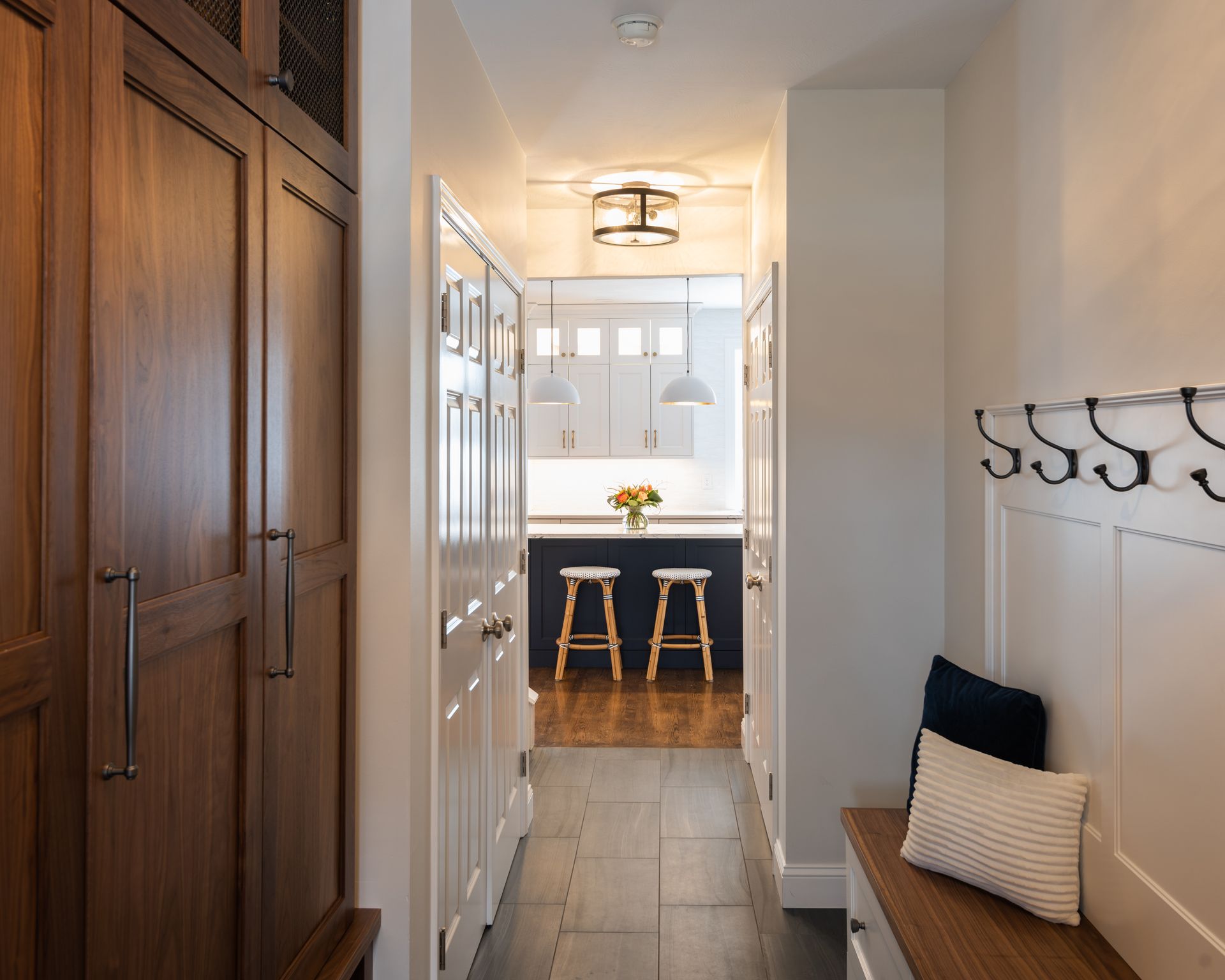 Narrow hallway with wooden cabinets, bench, coat hooks, and a bright kitchen with stools at the end.