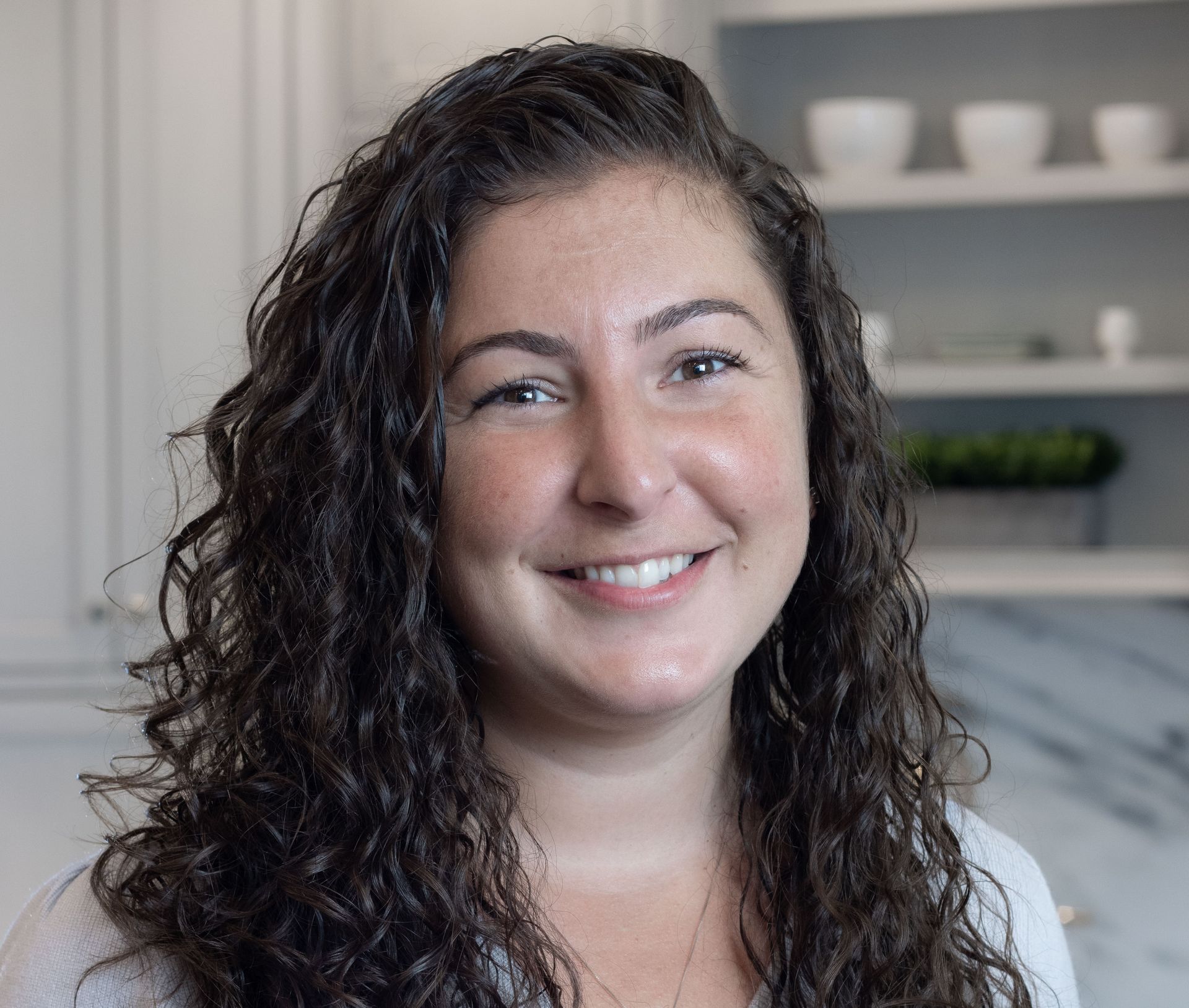 Smiling person with curly dark hair in a gray top, standing indoors near shelves.