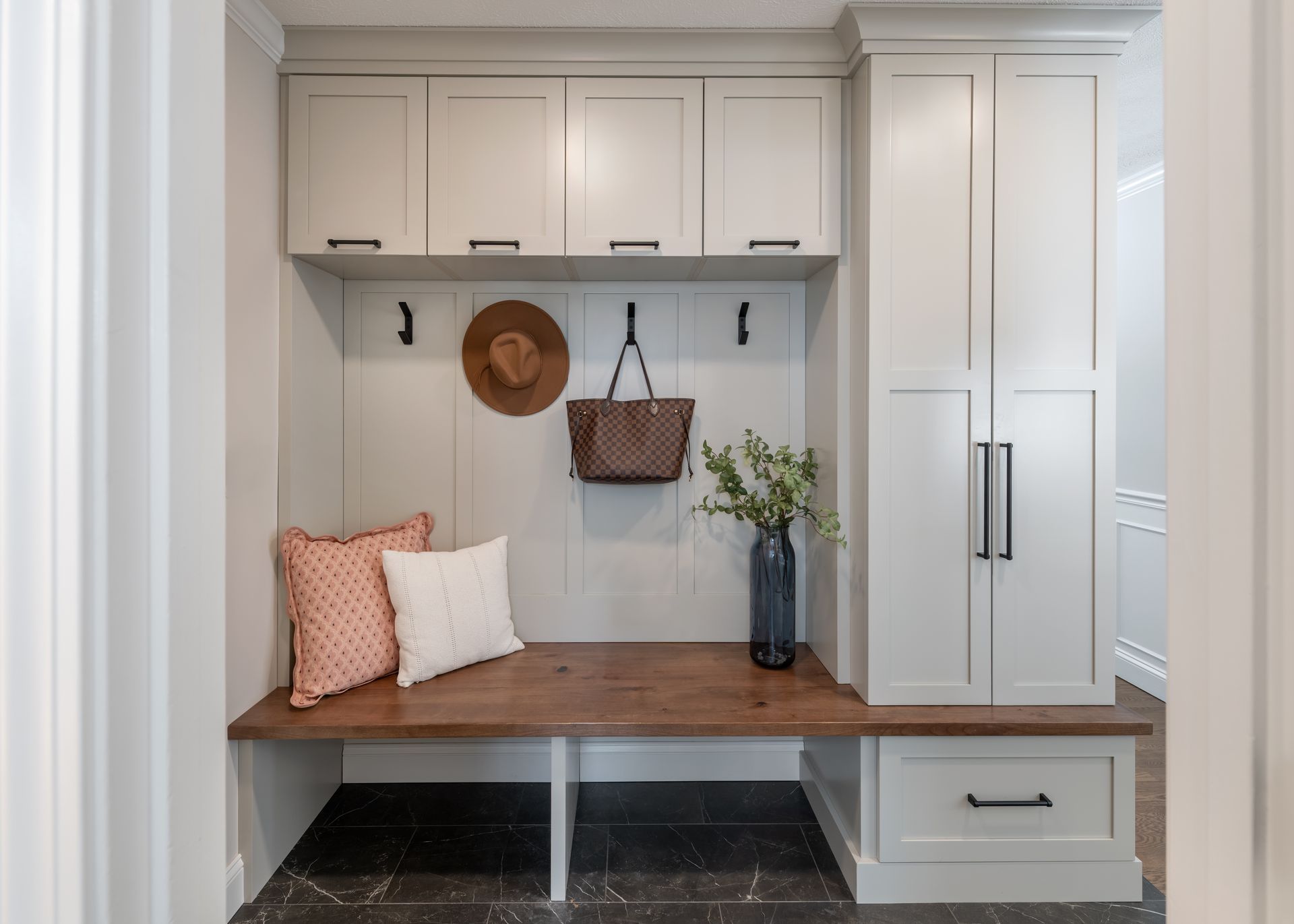 Built-in mudroom bench with hooks, cubbies, cushions, and a tall cabinet in a light gray entryway.