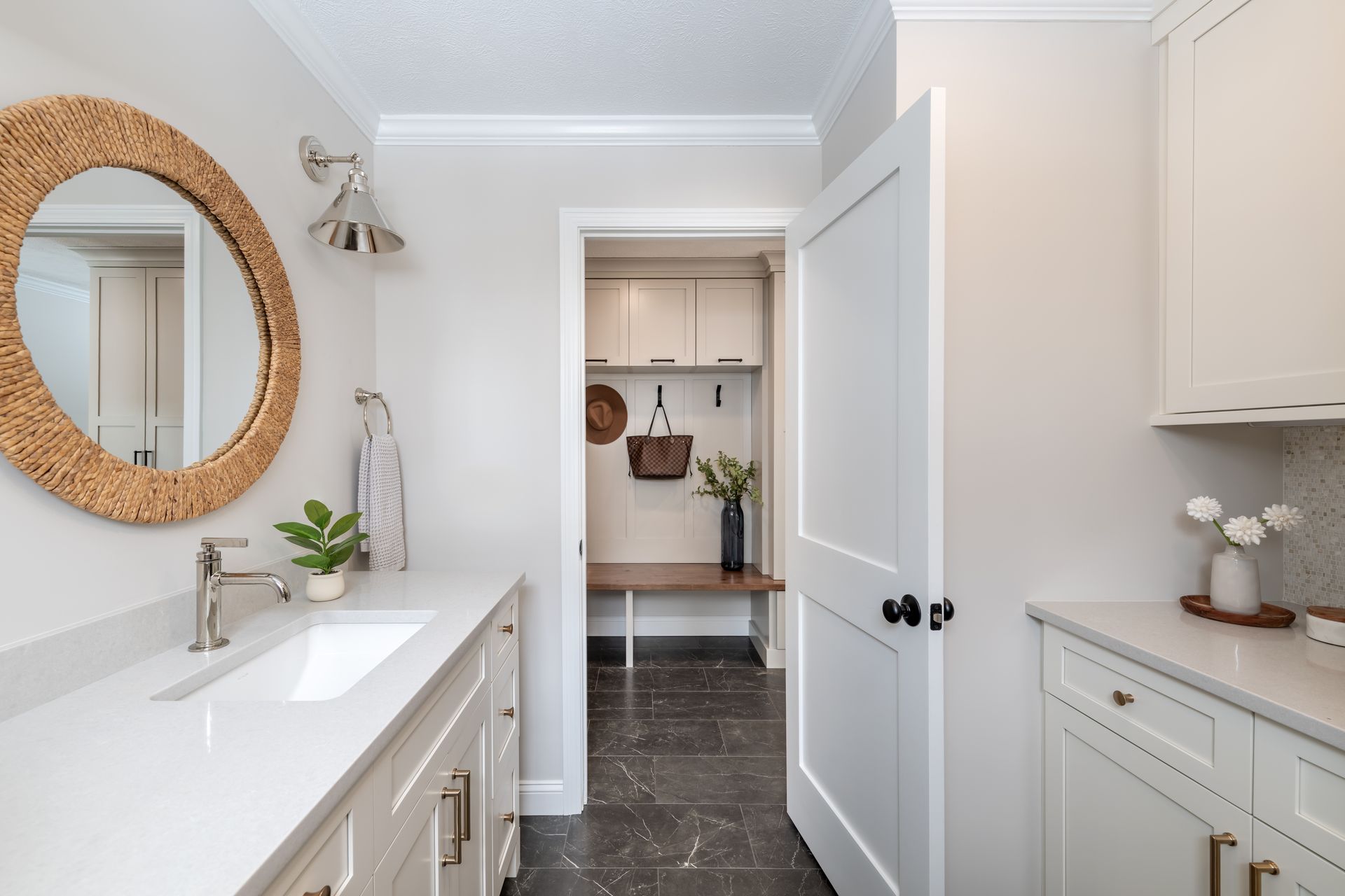 Bright bathroom with white vanity, round wicker mirror, and open door to a small kitchen area.