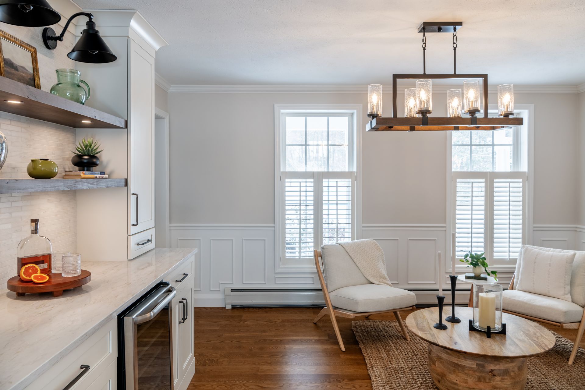 Bright living room with white wainscoting, wood floors, chandelier, and a cozy chair and rug by tall windows