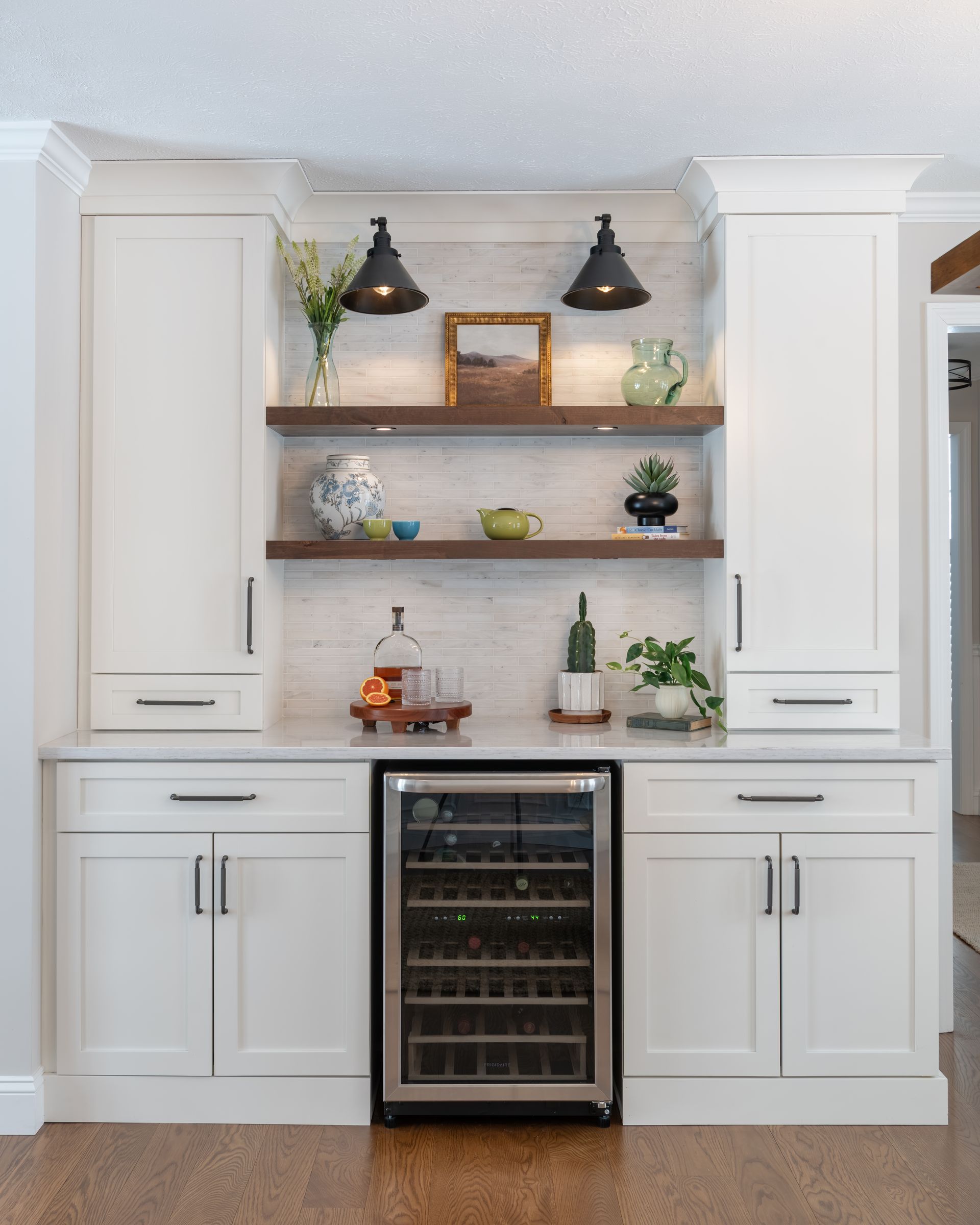 White kitchen bar with open shelves, pendant lights, and a wine fridge below countertops.