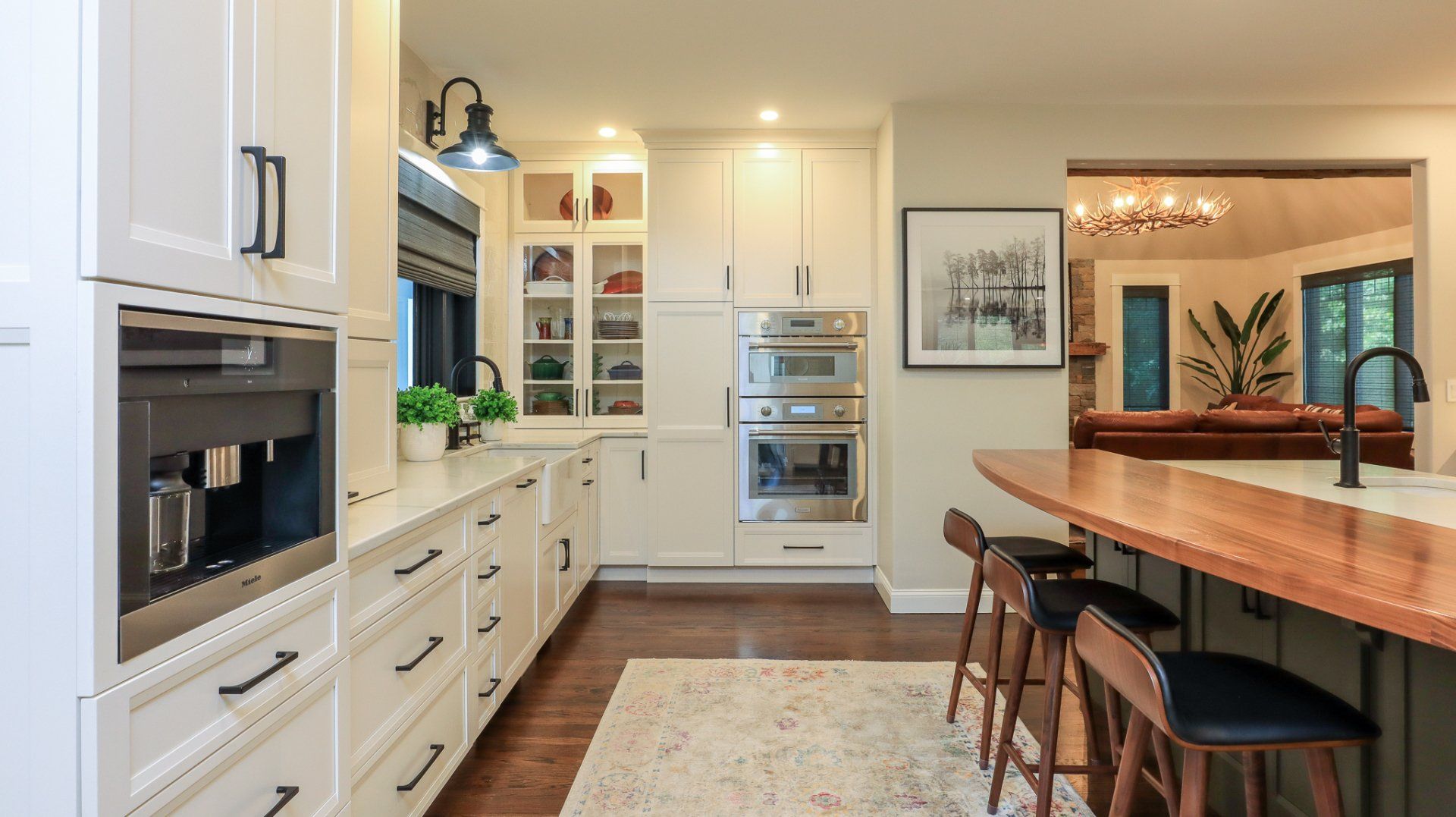 Bright modern kitchen with white cabinets, built-in ovens, wood island seating, and a dining area beyond.