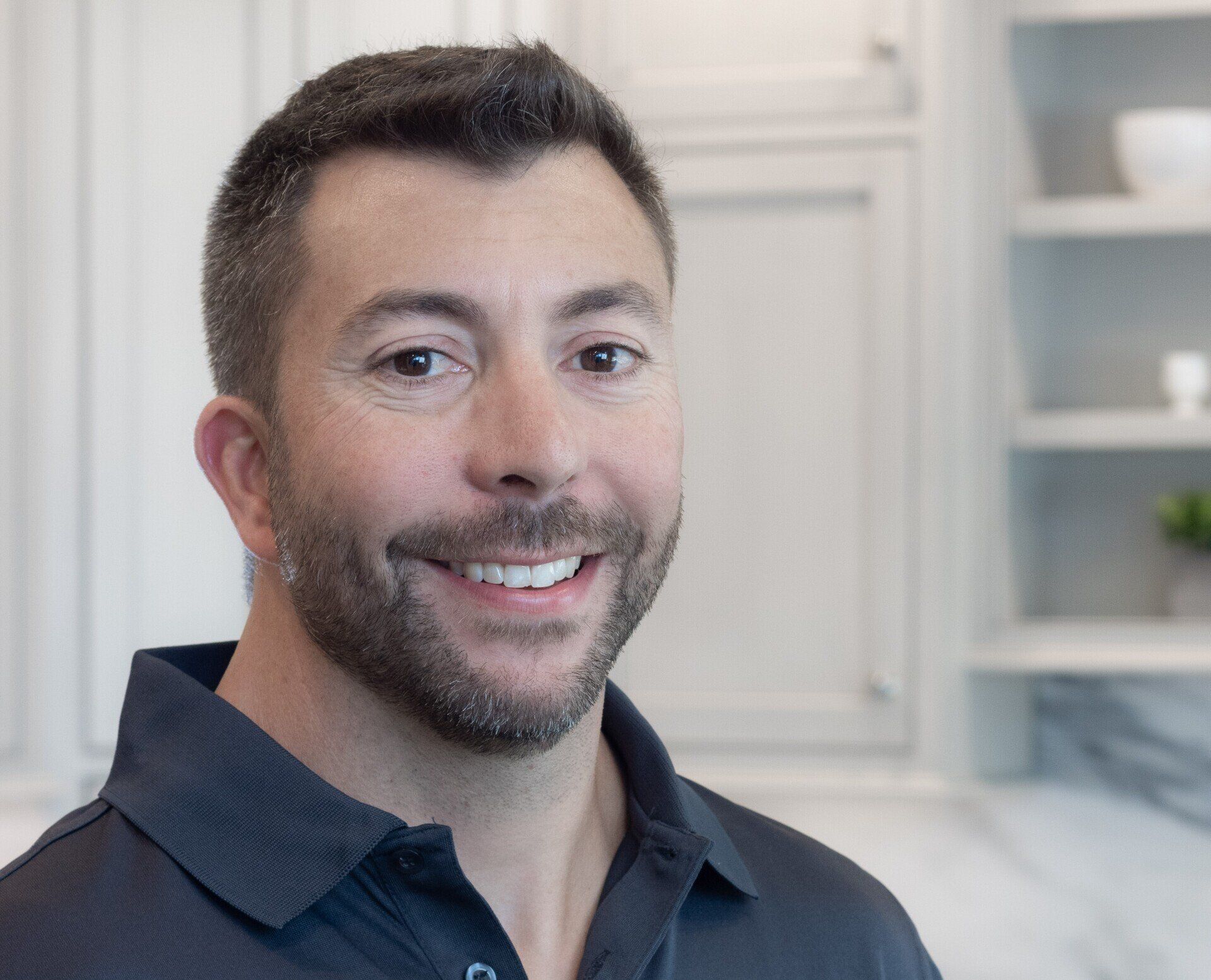 Smiling man in a dark polo shirt indoors, with white shelves in the background
