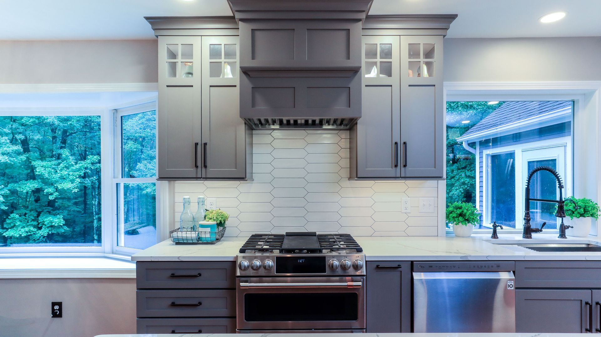 Modern kitchen with gray cabinets, stainless steel stove and dishwasher, and large windows overlooking greenery
