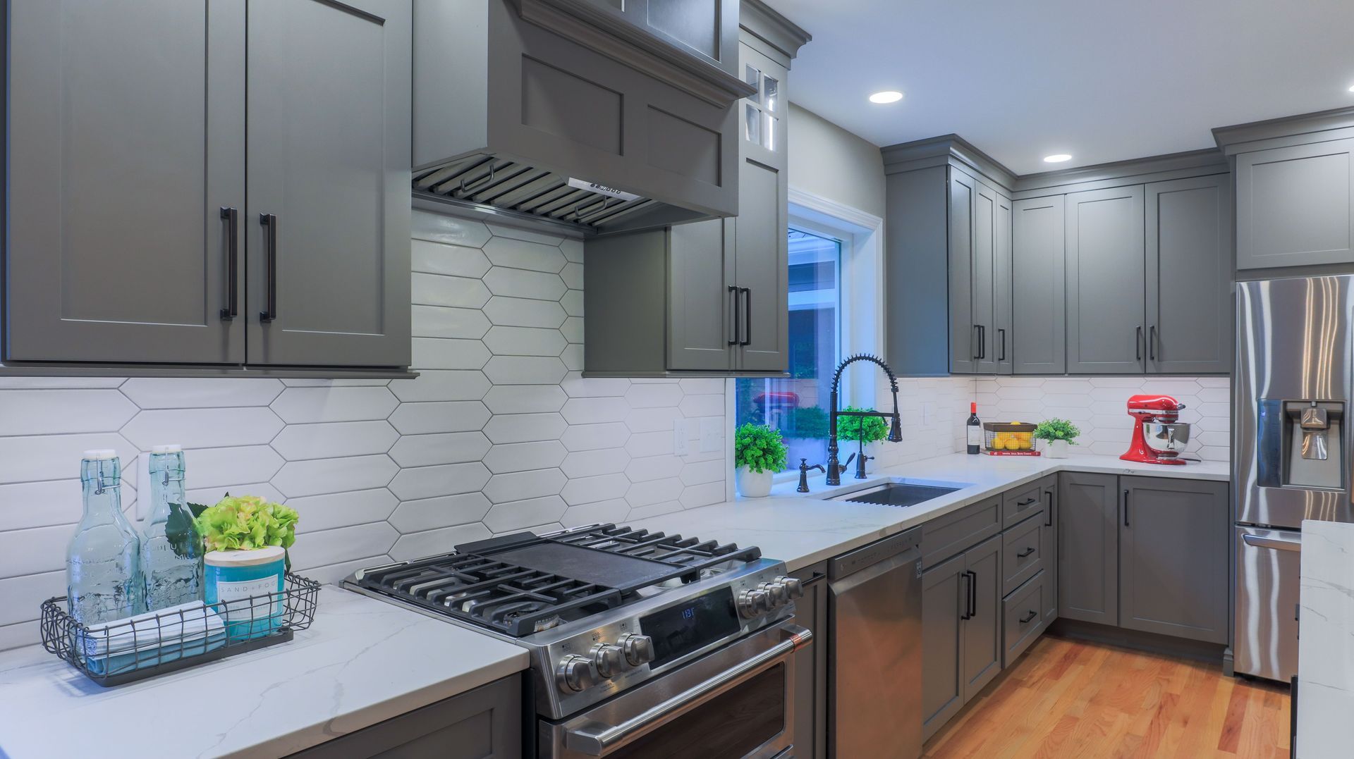 Modern kitchen with gray cabinets, white backsplash, stainless stove, and a sink by a bright window.