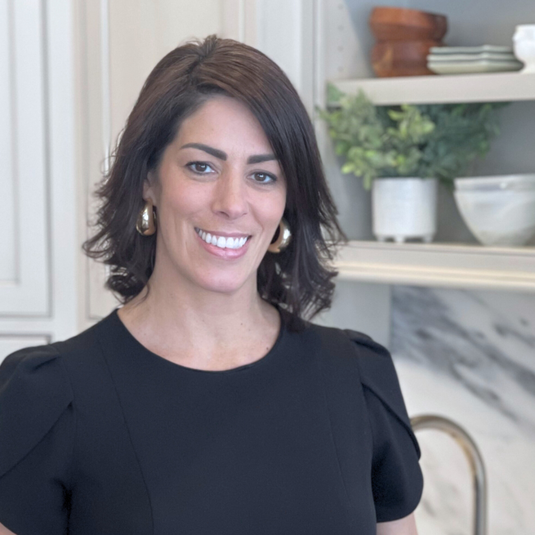 Smiling woman in a black top standing in a bright kitchen near marble counters and shelves