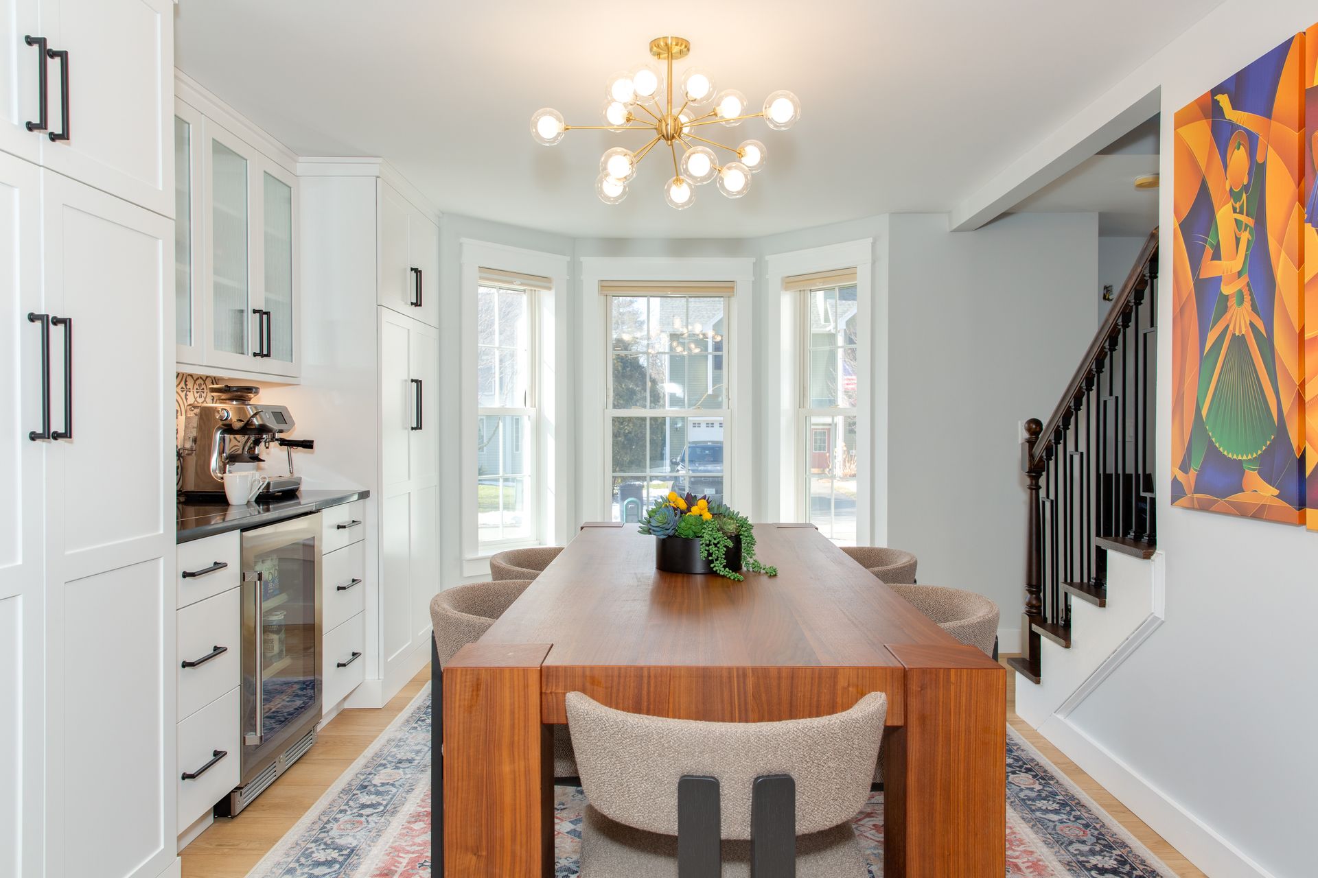 Bright modern dining room with long wooden table, chandelier, white cabinets, and colorful wall art