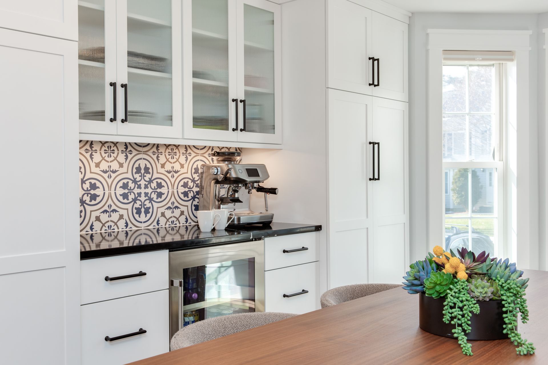 Bright white kitchen with blue patterned backsplash, glass cabinets, and a wooden table with flowers