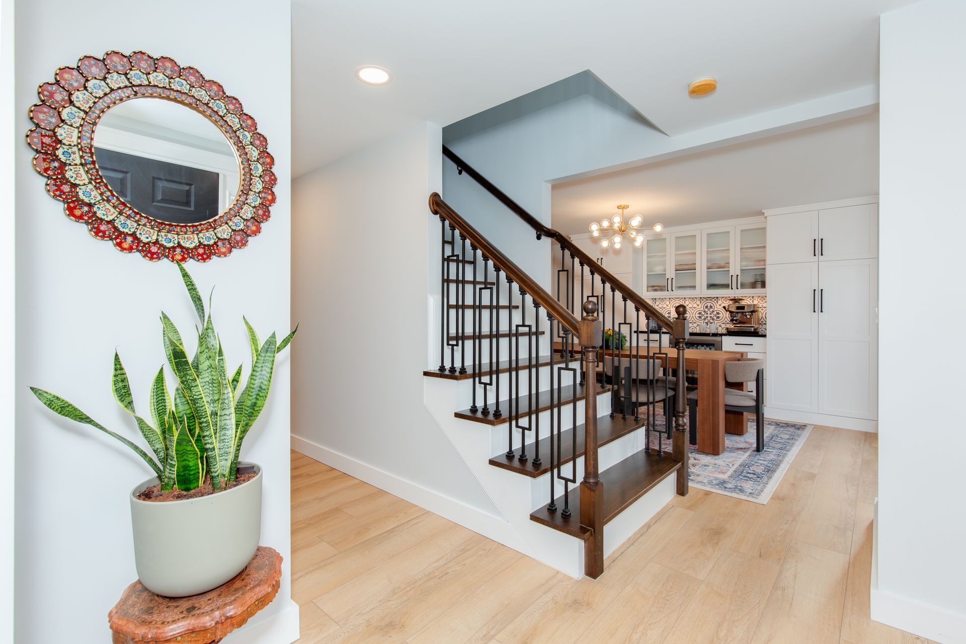 Bright foyer with staircase, wood floors, potted plant, and decorative mirror leading to a dining area