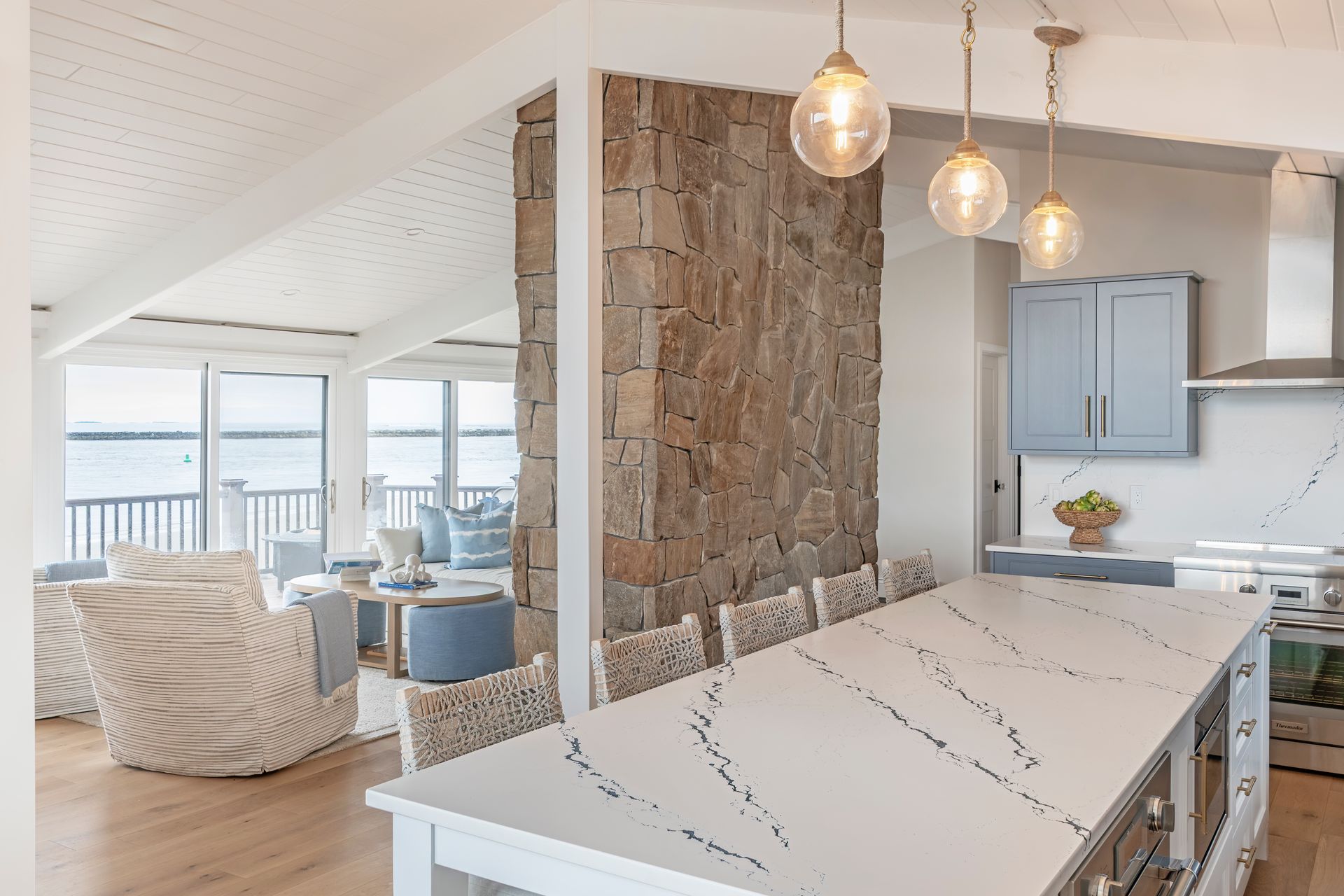 Bright kitchen island with pendant lights beside a stone accent wall and light-filled living area