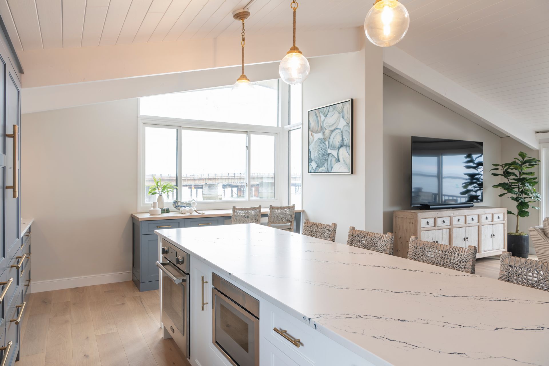 Bright modern kitchen with white island, pendant lights, and view into a living room with TV and sofa