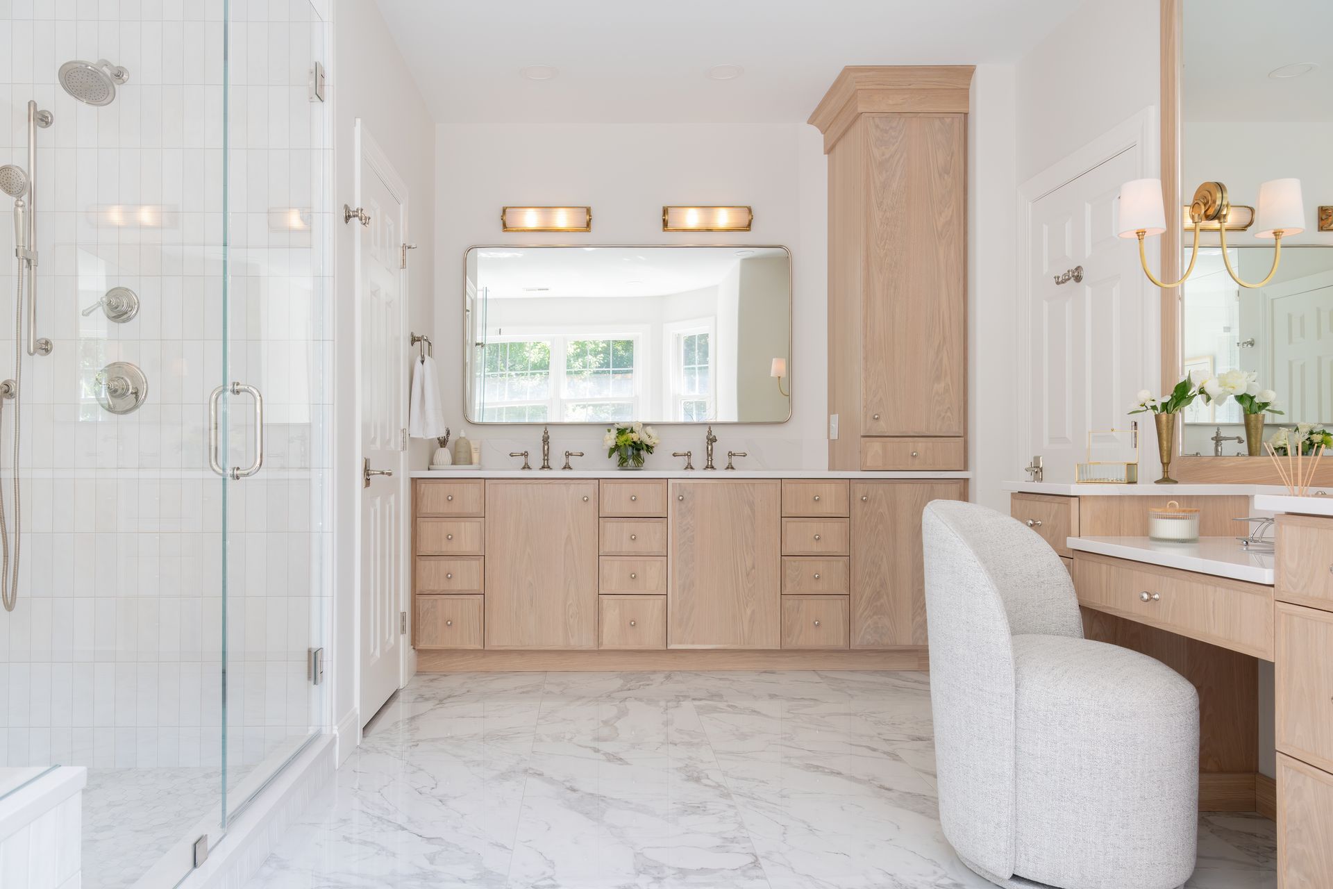 Bright luxury bathroom with white marble floors, wood vanities, large mirror, and glass shower.
