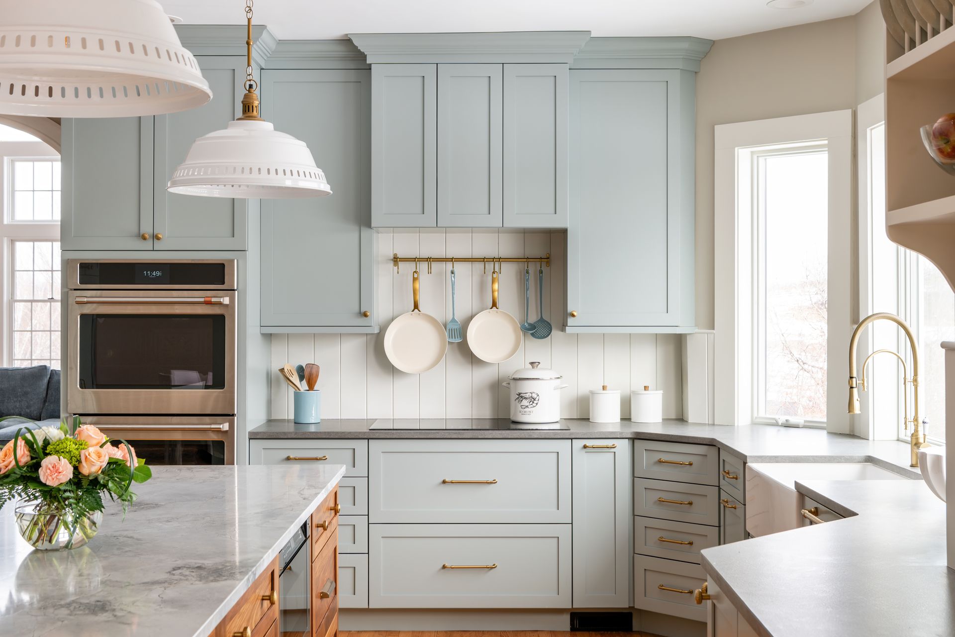 Bright white kitchen with pale blue cabinets, marble counters, and brass accents by a window