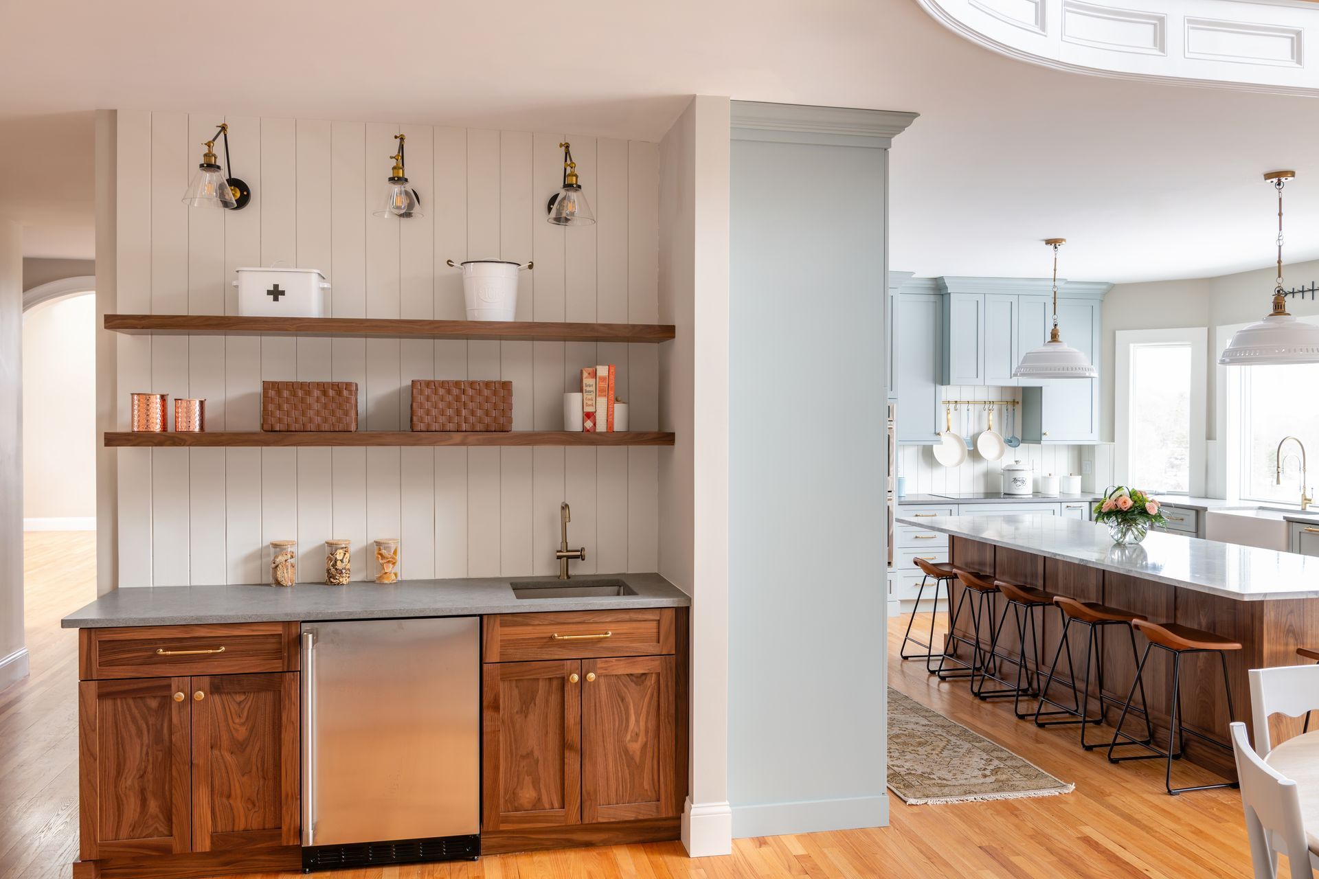 Built-in bar nook with shelves and wood cabinets beside a bright kitchen with island and stools