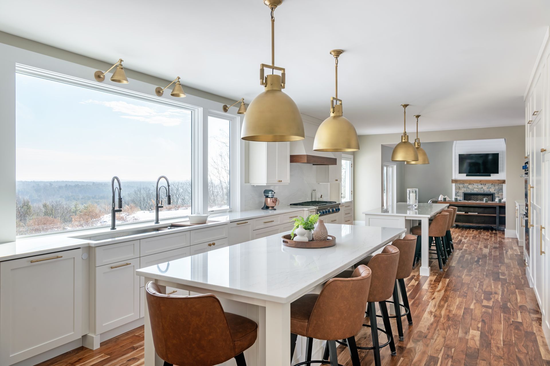 Bright modern kitchen with white island, tan stools, brass pendant lights, and large windows overlooking water