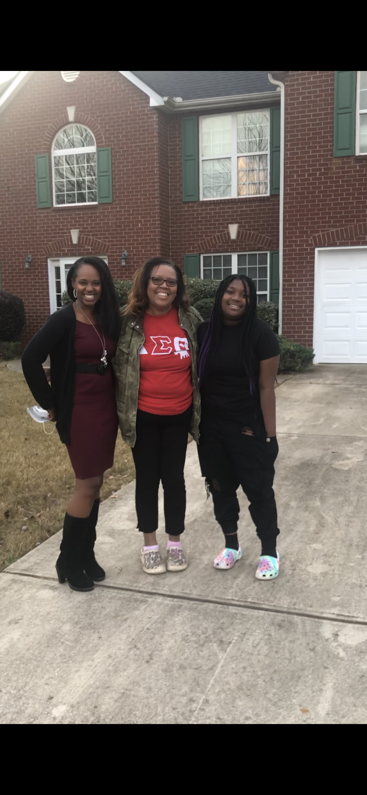 Three people stand on a driveway in front of a brick house. One wears a maroon dress, another red shirt, and the last, a black outfit.