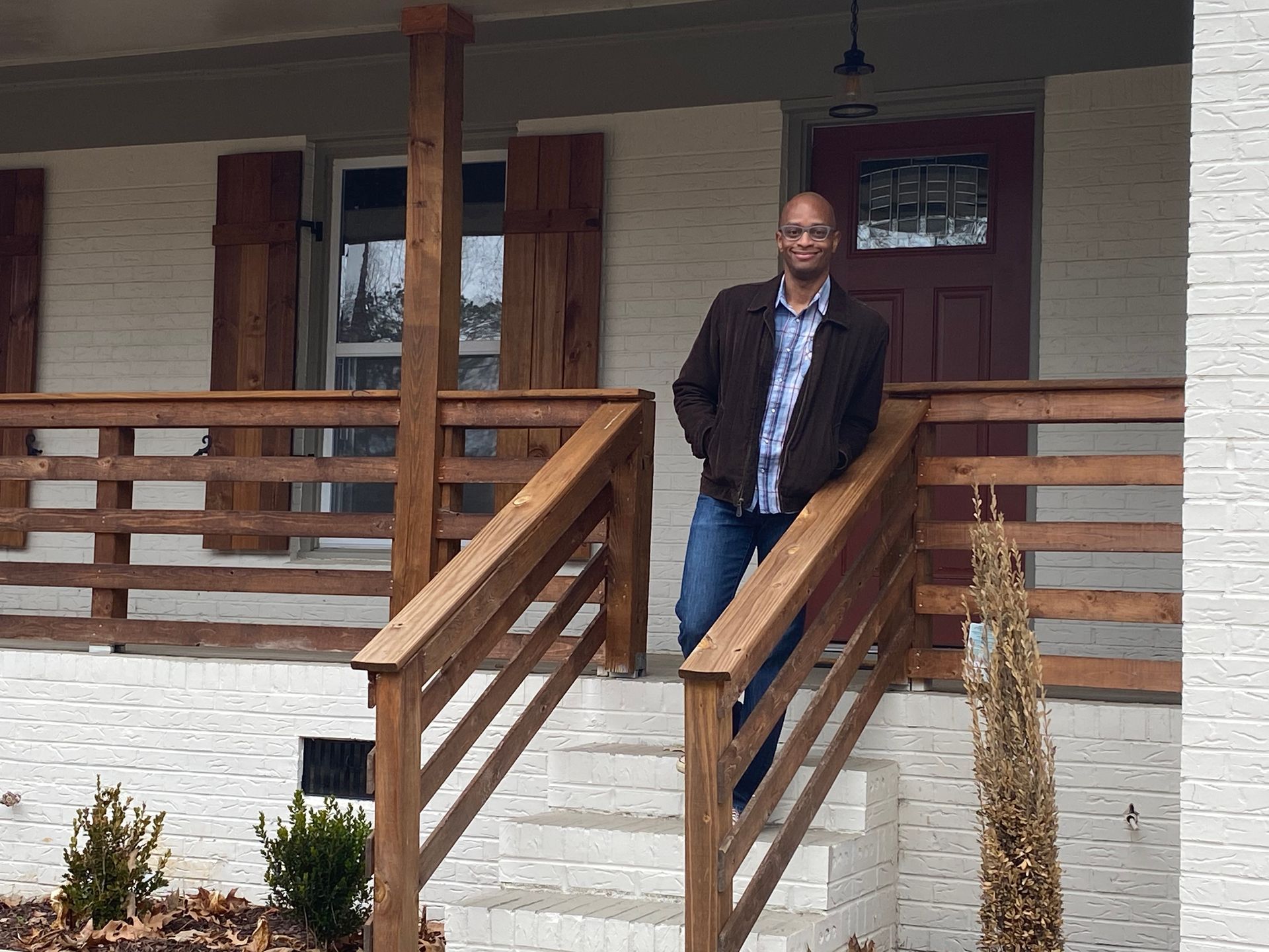 Man standing on the porch of a house with white brick exterior, wearing a brown jacket, hands in pockets.