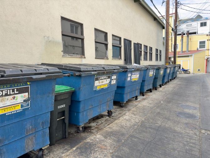 Row of blue and black dumpsters along a narrow alley beside a beige building